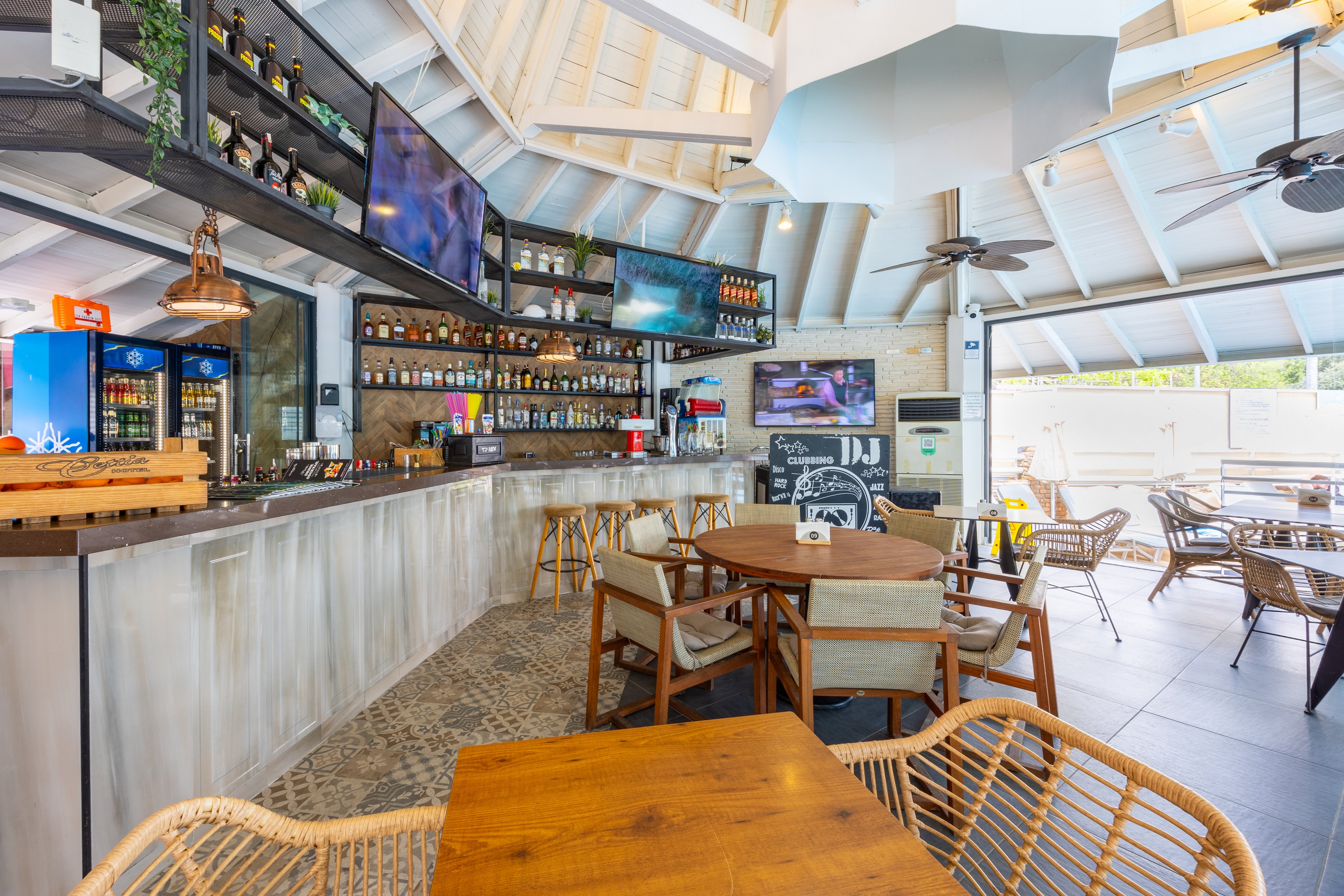 A contemporary bar interior features a long counter with a variety of bottles and glassware neatly arranged on shelves. Multiple flat-screen TVs are mounted above the bar, and a chalkboard menu is visible on the wall. The setting includes wooden tables and wicker chairs, with natural light streaming in through large windows. The overall palette is neutral with light wood and white tones, creating a relaxed and inviting atmosphere.