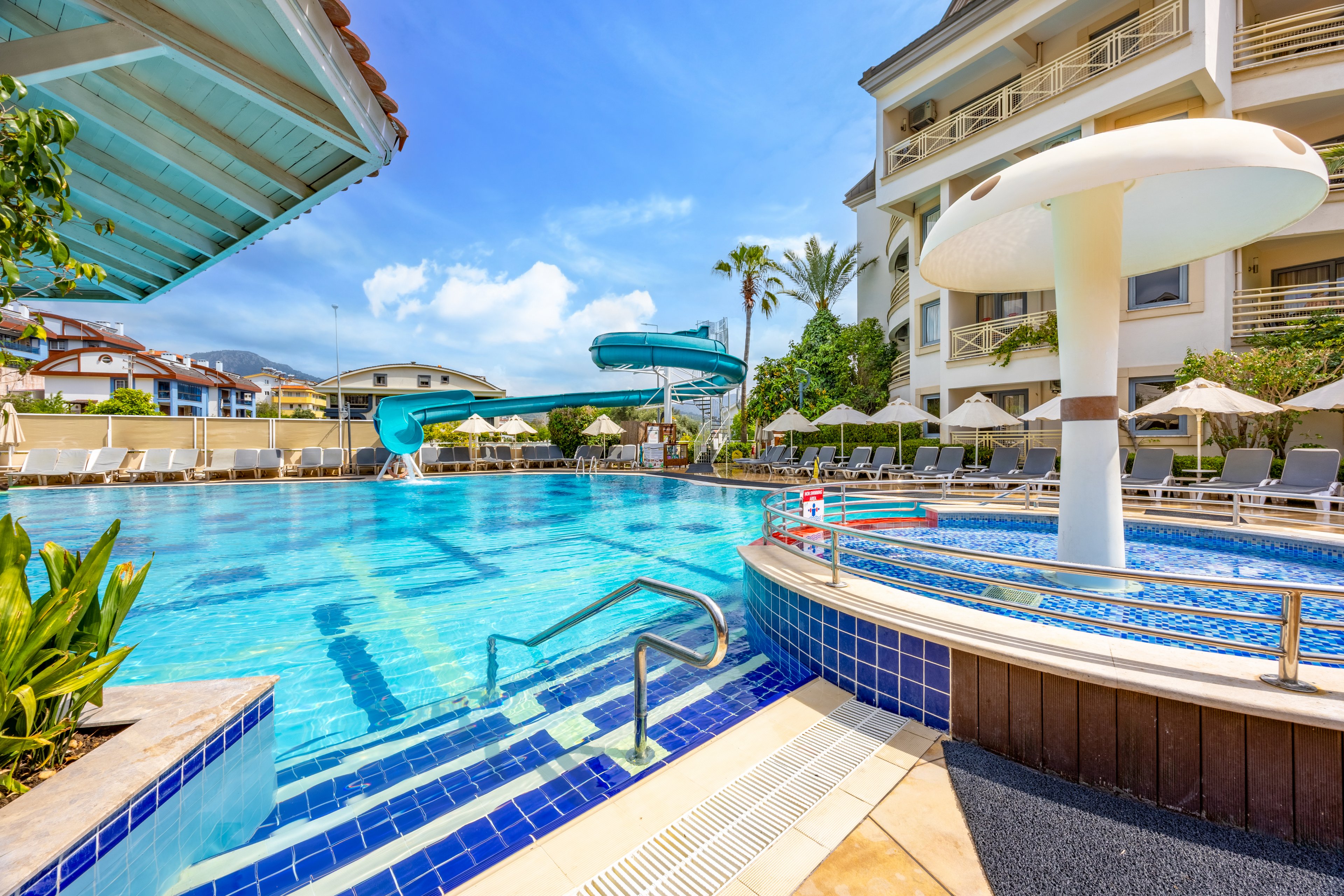 A large outdoor swimming pool is featured at a resort, with a prominent blue waterslide and a white mushroom-shaped umbrella fountain. The pool area is surrounded by lounge chairs and sun umbrellas, with a modern hotel building in the background. The setting is bright and sunny, with clear blue skies and palm trees adding to the vacation atmosphere. No visible people are present, emphasizing a calm and inviting mood.