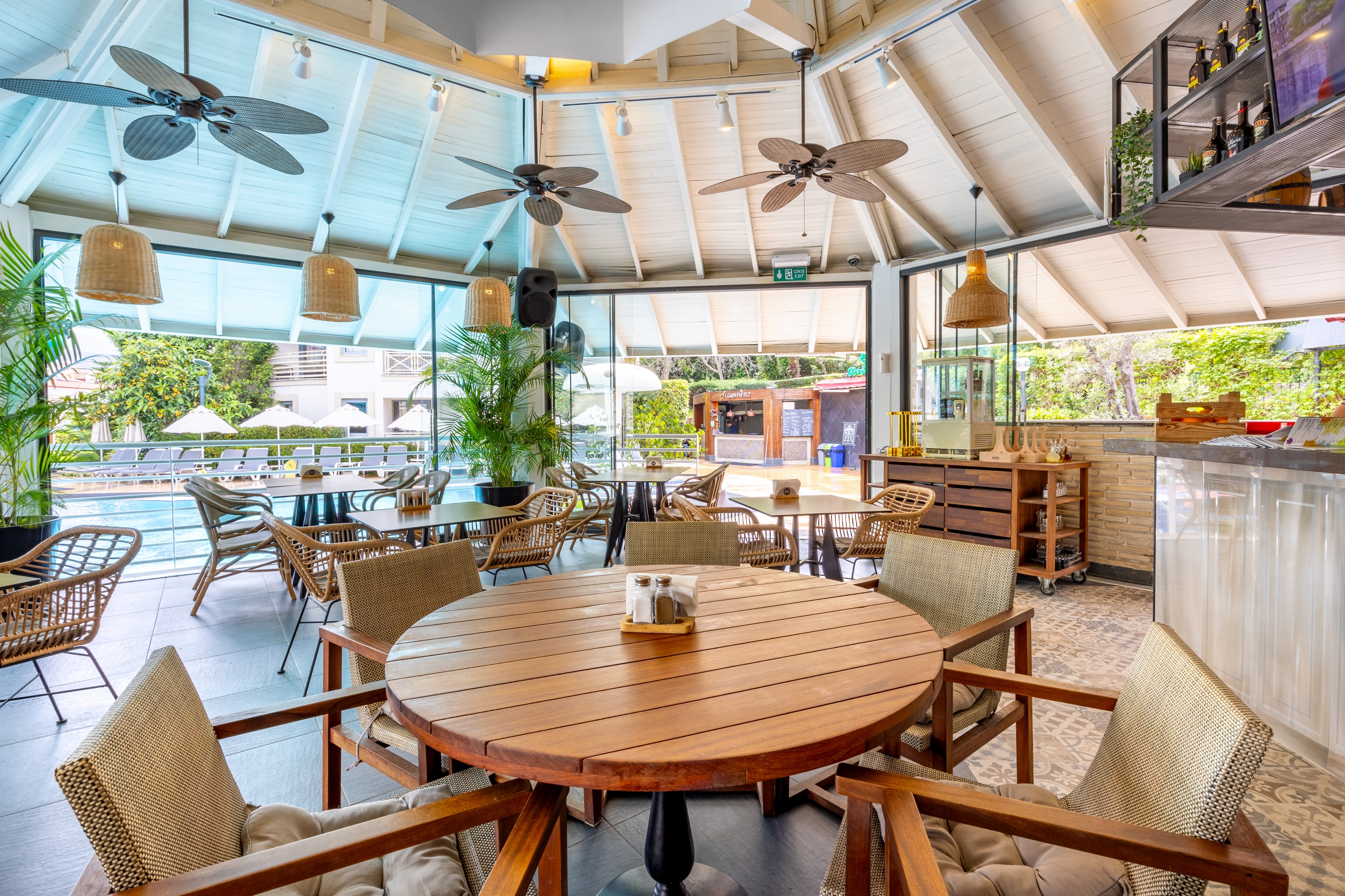 A spacious, sunlit restaurant features wooden tables and woven chairs arranged in a circular layout. Large windows and open walls provide a clear view of the outdoor pool area and lush greenery. Ceiling fans, pendant lights, and natural decor elements create a relaxed, tropical atmosphere. The setting is casual and inviting, ideal for social gatherings or leisurely dining. No visible text or numbers are present in the image.
