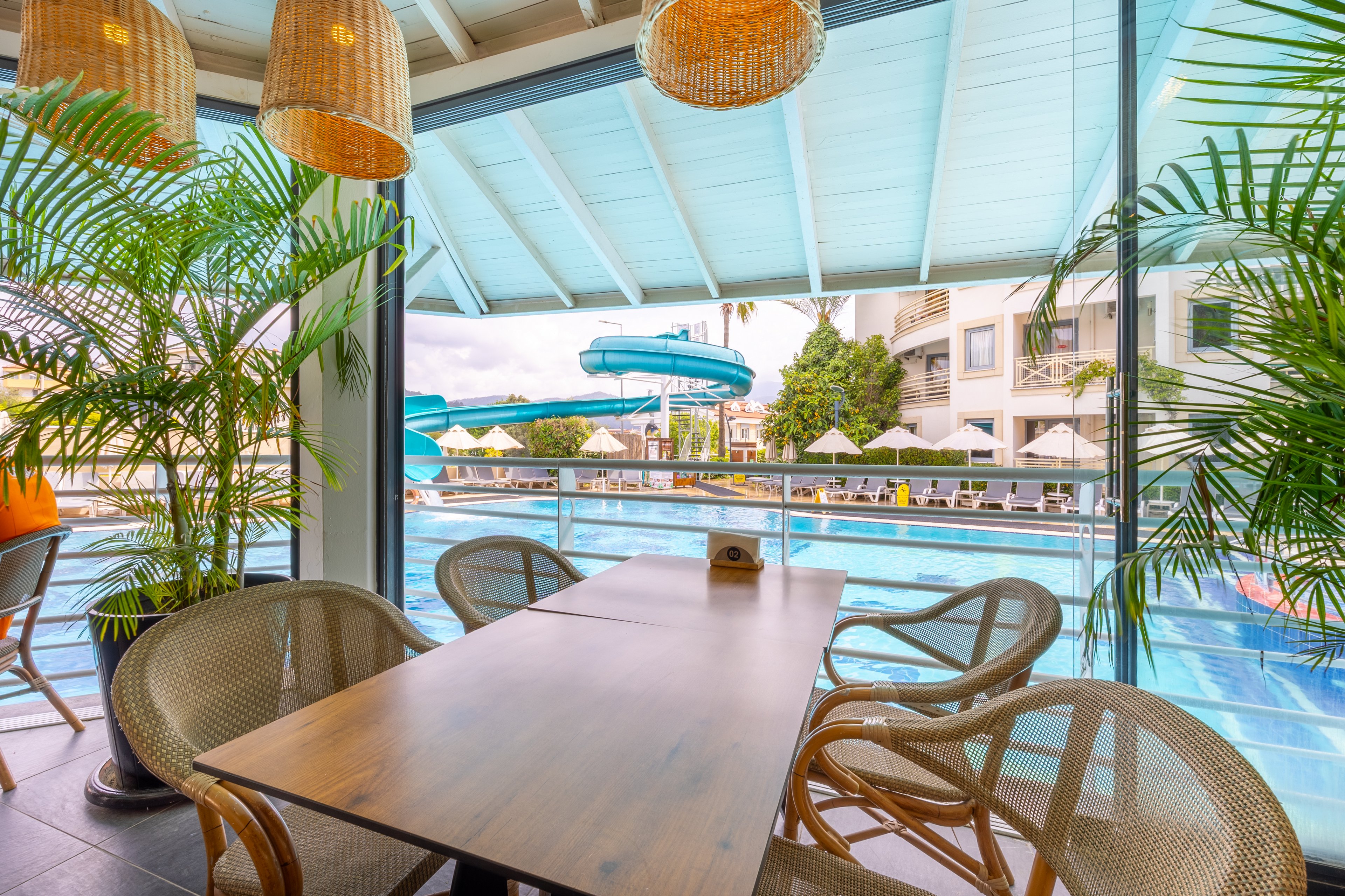 A sunlit dining area features a wooden table and wicker chairs, set beside large windows overlooking a vibrant outdoor pool. A blue waterslide curves into the pool, with resort buildings and umbrellas visible in the background. Lush green plants and woven pendant lights add a tropical, relaxed atmosphere. The setting is empty, emphasizing tranquility and leisure. No visible text or numbers are present in the image.
