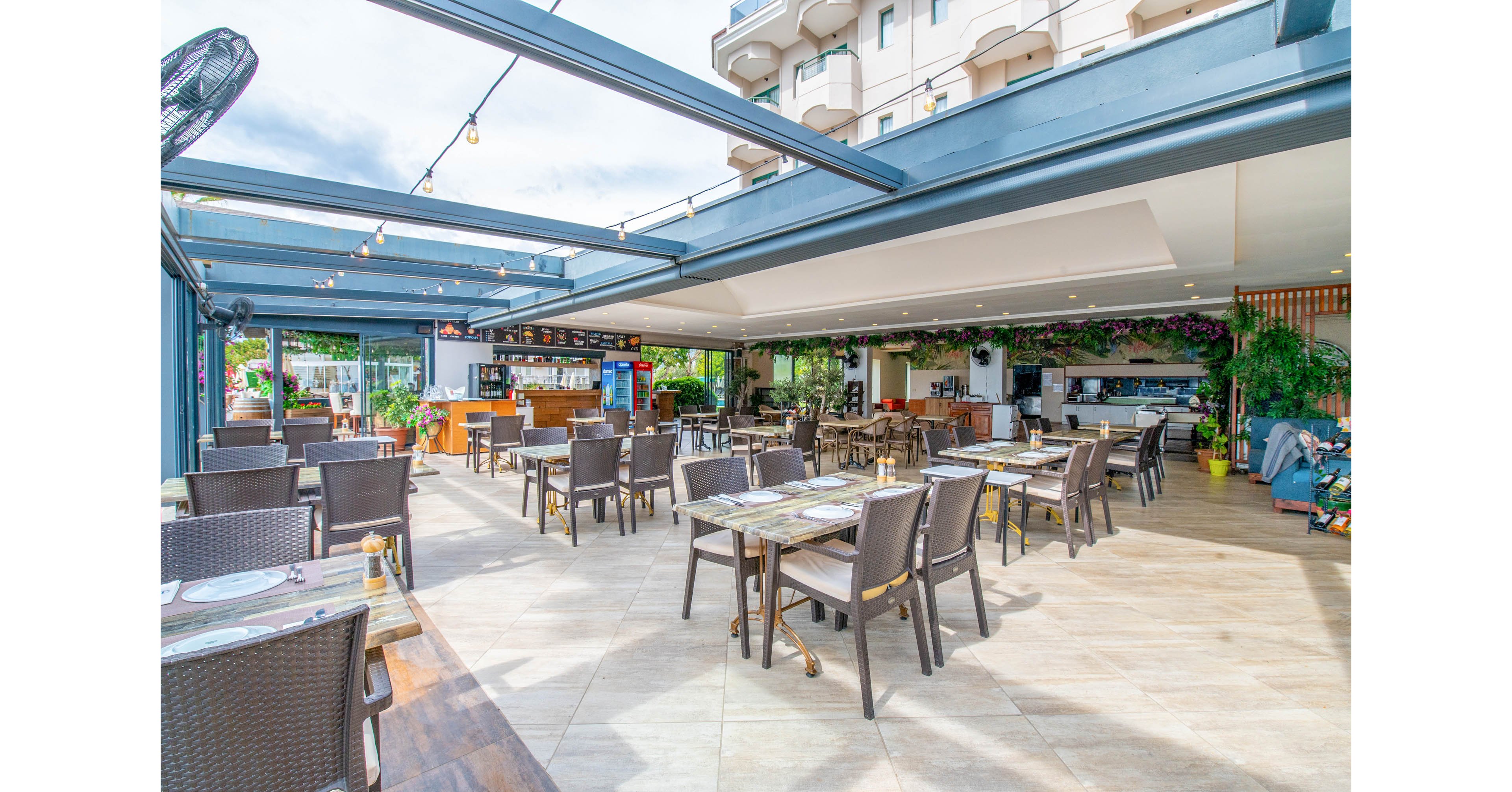 Spacious outdoor restaurant area with numerous tables and wicker chairs arranged on a tiled terrace. The setting appears to be part of a hotel or resort, with an open roof structure and bright daylight creating an airy atmosphere. A bar and food counter are visible in the background, along with plants and decorative lighting that add a welcoming touch. The space is currently empty of guests, emphasizing the layout and design of the dining area.