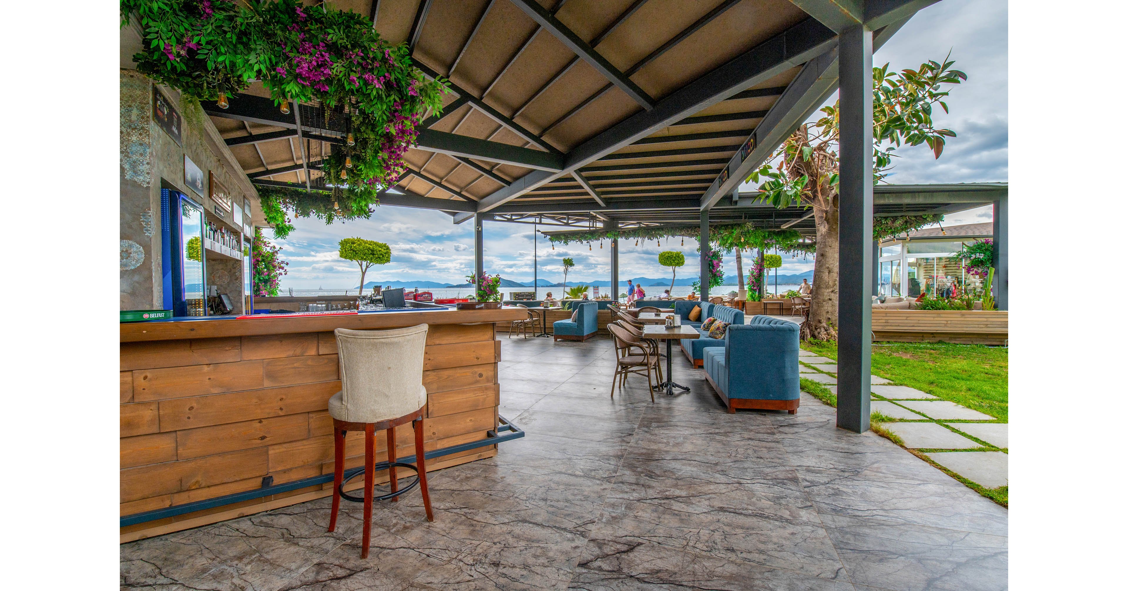 An open-air bar with wooden counter and hanging flowers overlooks a seaside promenade. Comfortable lounge chairs and tables are arranged under a modern metal roof structure. The polished stone floor leads toward a grassy area with trimmed trees and ocean views in the background. A few people can be seen in the distance near the water, adding a relaxed coastal atmosphere.