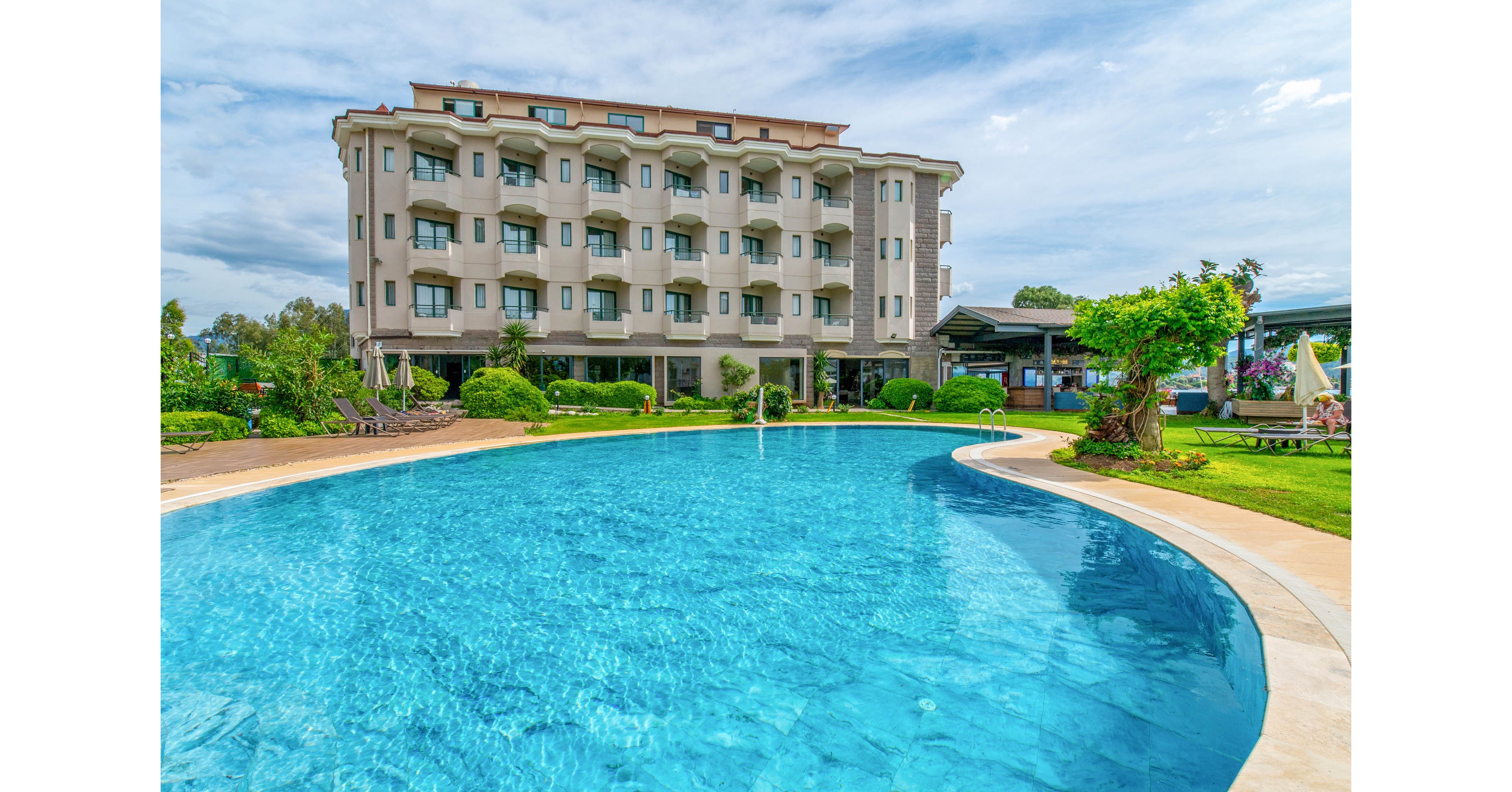 A multi-story resort hotel overlooks a large curved outdoor swimming pool. The bright blue water contrasts with the beige facade of the building and the green landscaped gardens surrounding the pool. Lounge chairs and umbrellas are visible near the poolside, suggesting a relaxing vacation setting. The sky is partly cloudy, adding a soft, natural backdrop to the tranquil scene.