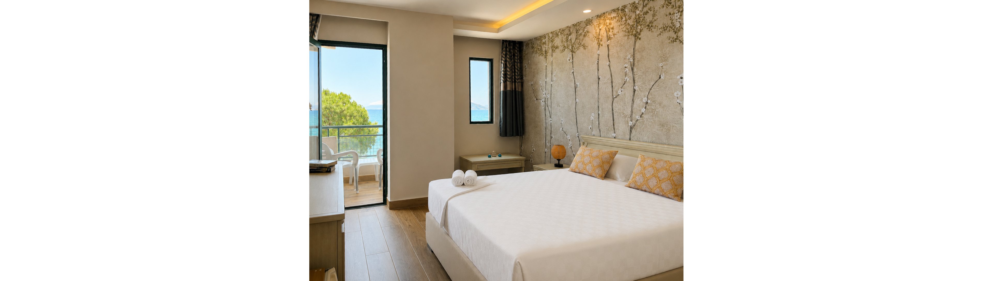 A bright, modern hotel bedroom features a large neatly made bed with two patterned pillows and rolled white towels placed on top. The room opens onto a balcony with chairs, overlooking a scenic sea and distant mountains. Warm wood flooring, soft neutral walls, and a decorative tree-patterned accent wall create a calm, contemporary atmosphere. Subtle lighting along the ceiling adds to the cozy, inviting feel of the space.