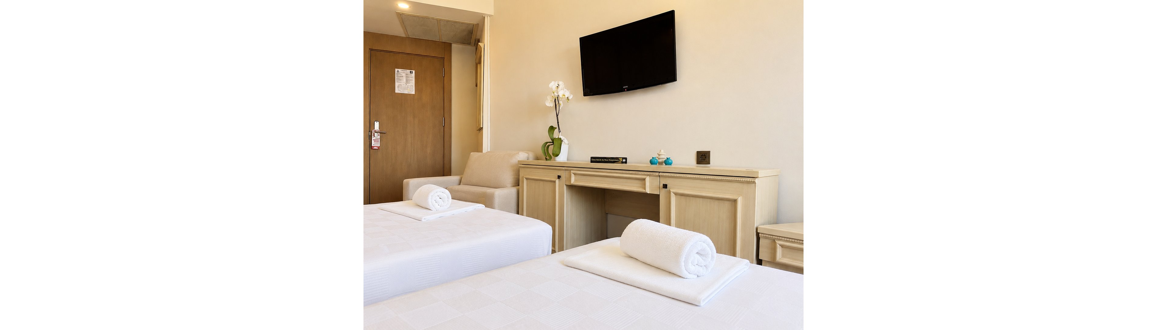A bright hotel room features two neatly made twin beds with rolled white towels placed at the foot. A light wooden desk runs along the wall, holding a potted orchid, a book, and small decorative items. Above the desk, a flat-screen television is mounted on the beige wall. A cushioned armchair and a wooden door with a keycard lock complete the clean, contemporary interior.