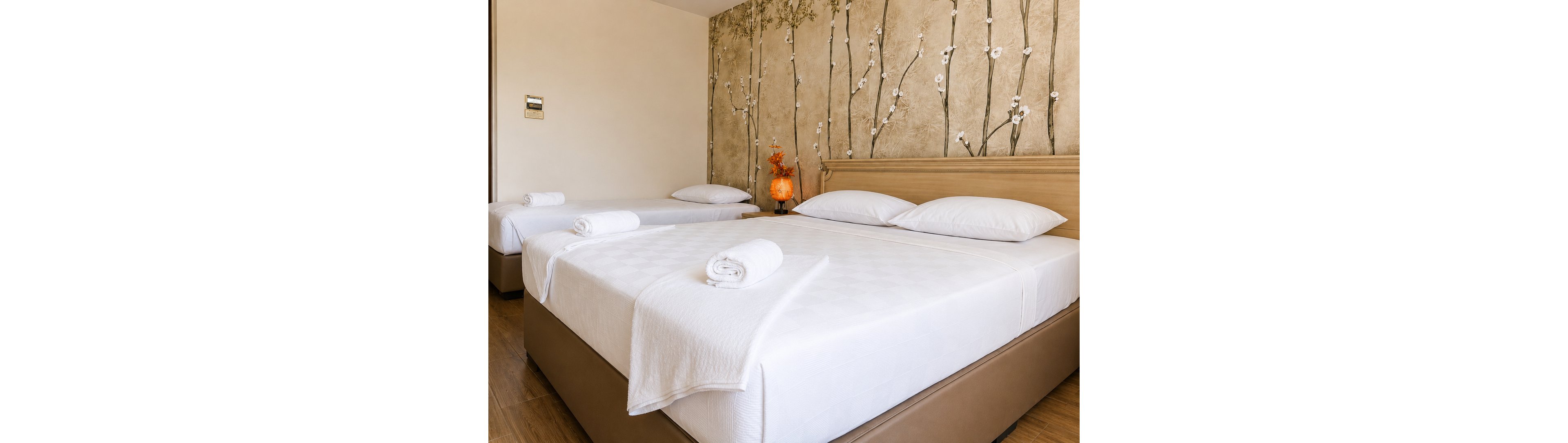 A neatly arranged hotel room features a large central bed flanked by two smaller beds, all topped with white linens and rolled towels. The room has a warm wooden floor and a light-colored headboard running along the wall. A decorative wall mural with delicate branches and blossoms adds an elegant touch behind the beds. A small bedside table holds an orange floral arrangement and a glowing lamp, creating a cozy and inviting atmosphere.