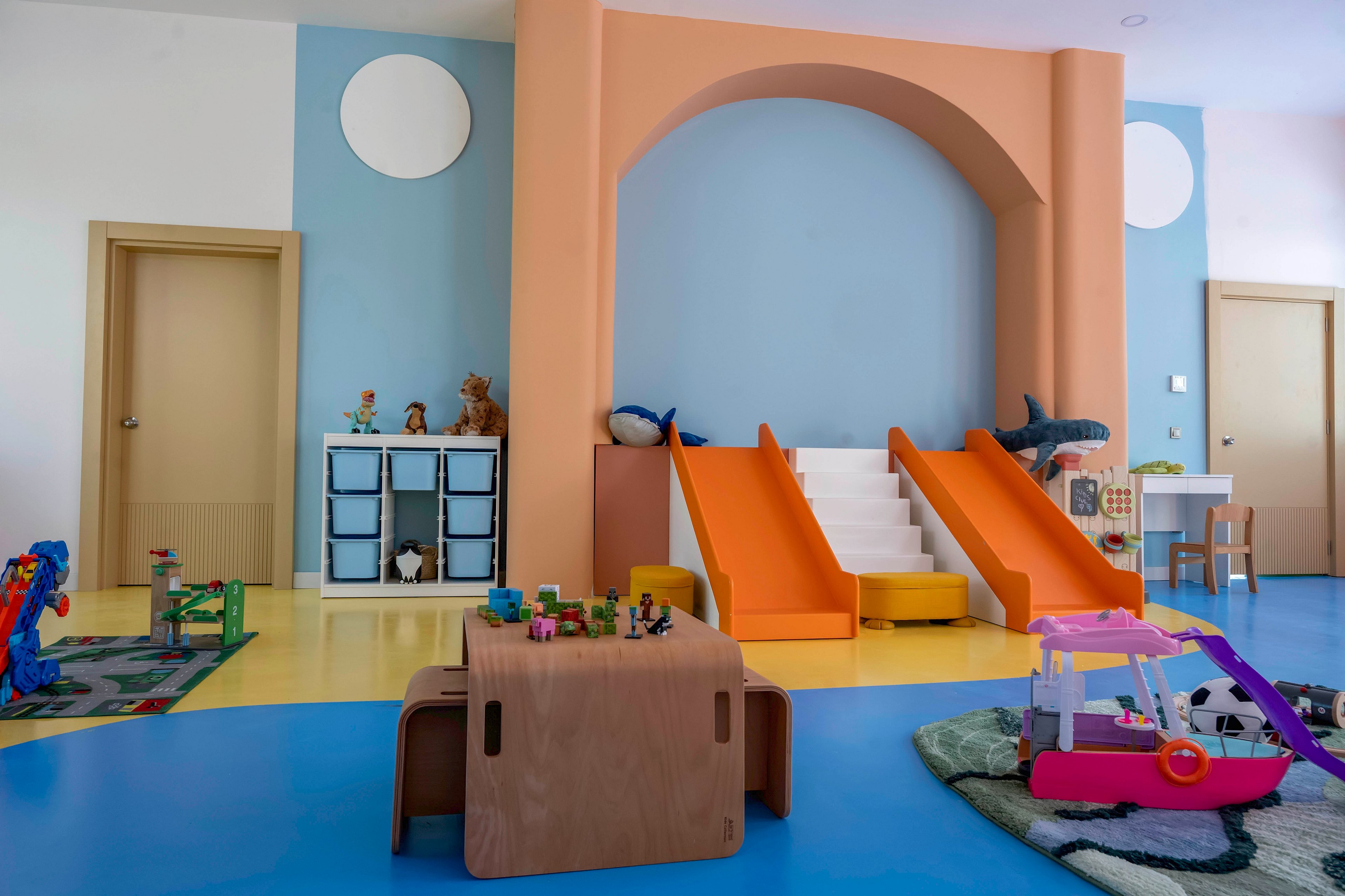 A bright indoor playroom features two orange slides with white steps as the central attraction. The room has blue and yellow flooring, pastel-colored walls, and multiple doors. Various toys, including building blocks, figurines, and playsets, are scattered on low tables and rugs. Shelves with stuffed animals and storage bins line the wall, creating a playful and organized environment for children.