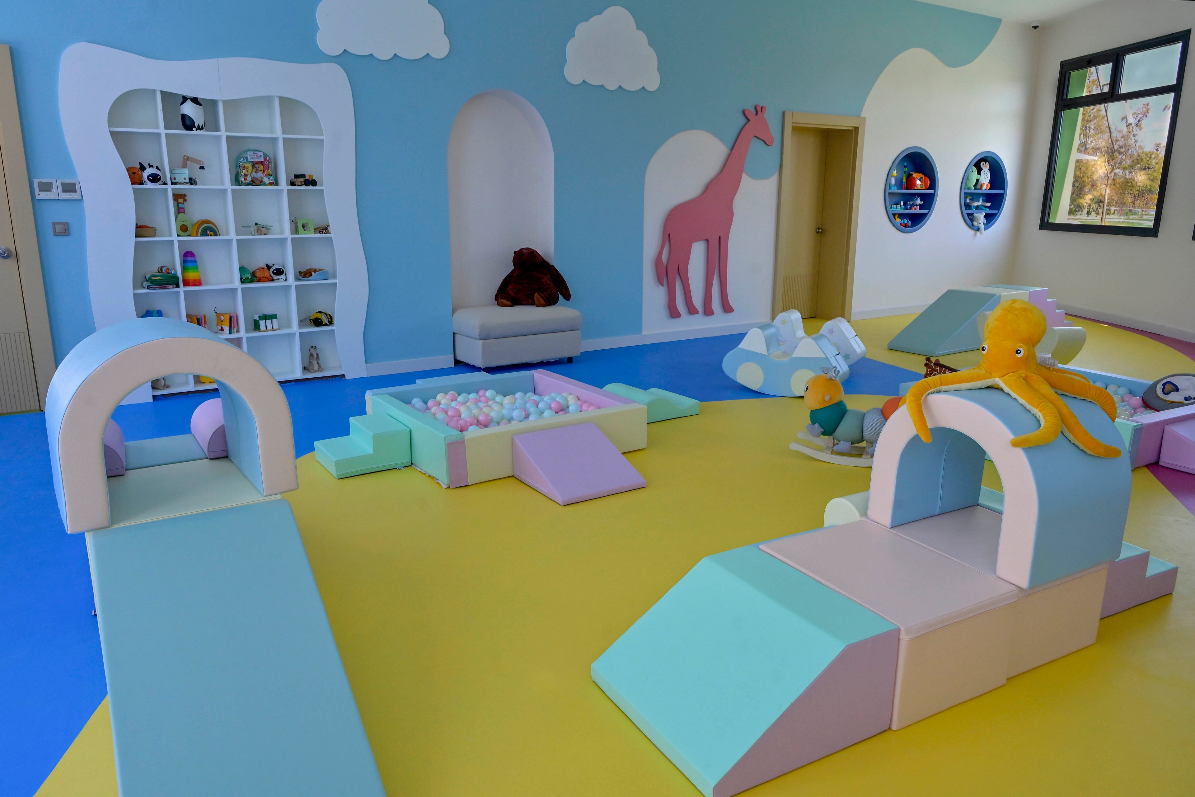 A bright indoor playroom features soft foam structures and a small ball pit in pastel colors. The room is decorated with cloud shapes, a pink giraffe mural, and playful wall cutouts. Shelves with stuffed toys and a cushioned chair sit against the back wall. Large windows and round wall openings bring in natural light, creating a cheerful and safe environment for young children.