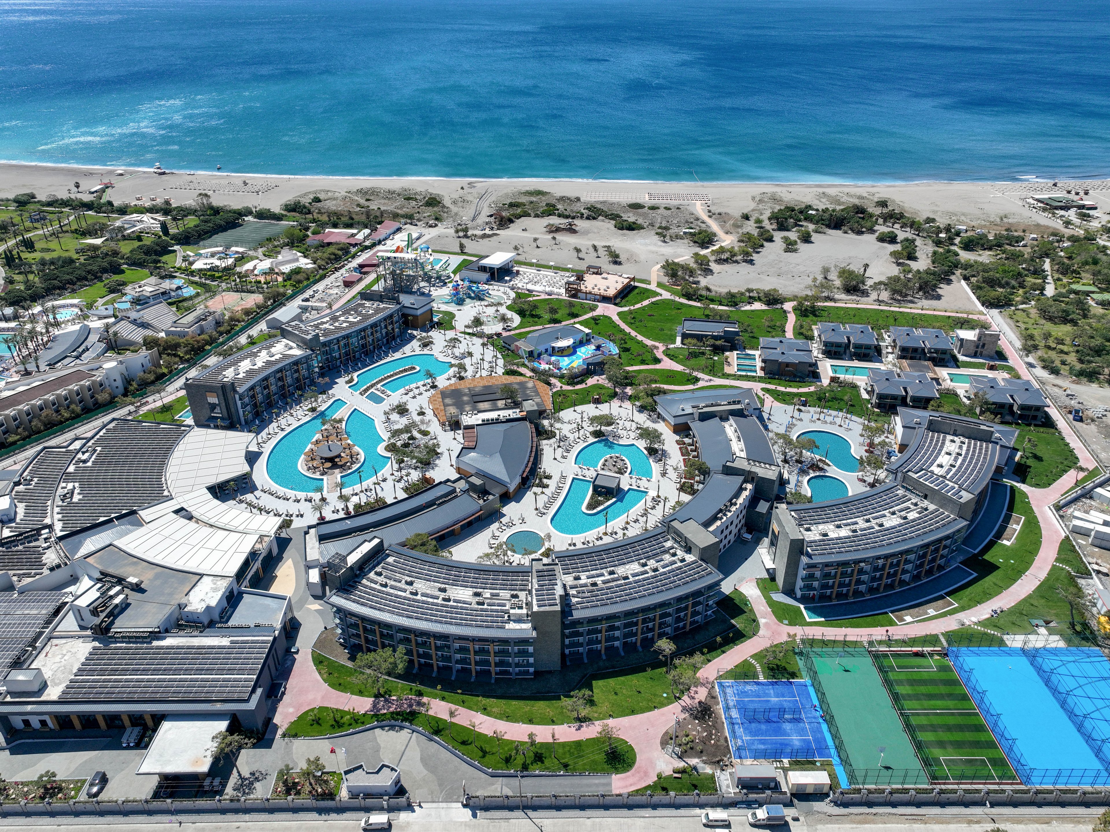 A large coastal resort complex is captured from an aerial perspective beside a bright blue sea. Curved hotel buildings surround multiple outdoor swimming pools and sunbathing areas. Sports facilities, including tennis courts and other recreational spaces, are visible near the bottom of the image. Sandy beach, scattered vegetation, and walking paths extend toward the shoreline, emphasizing the vacation setting.