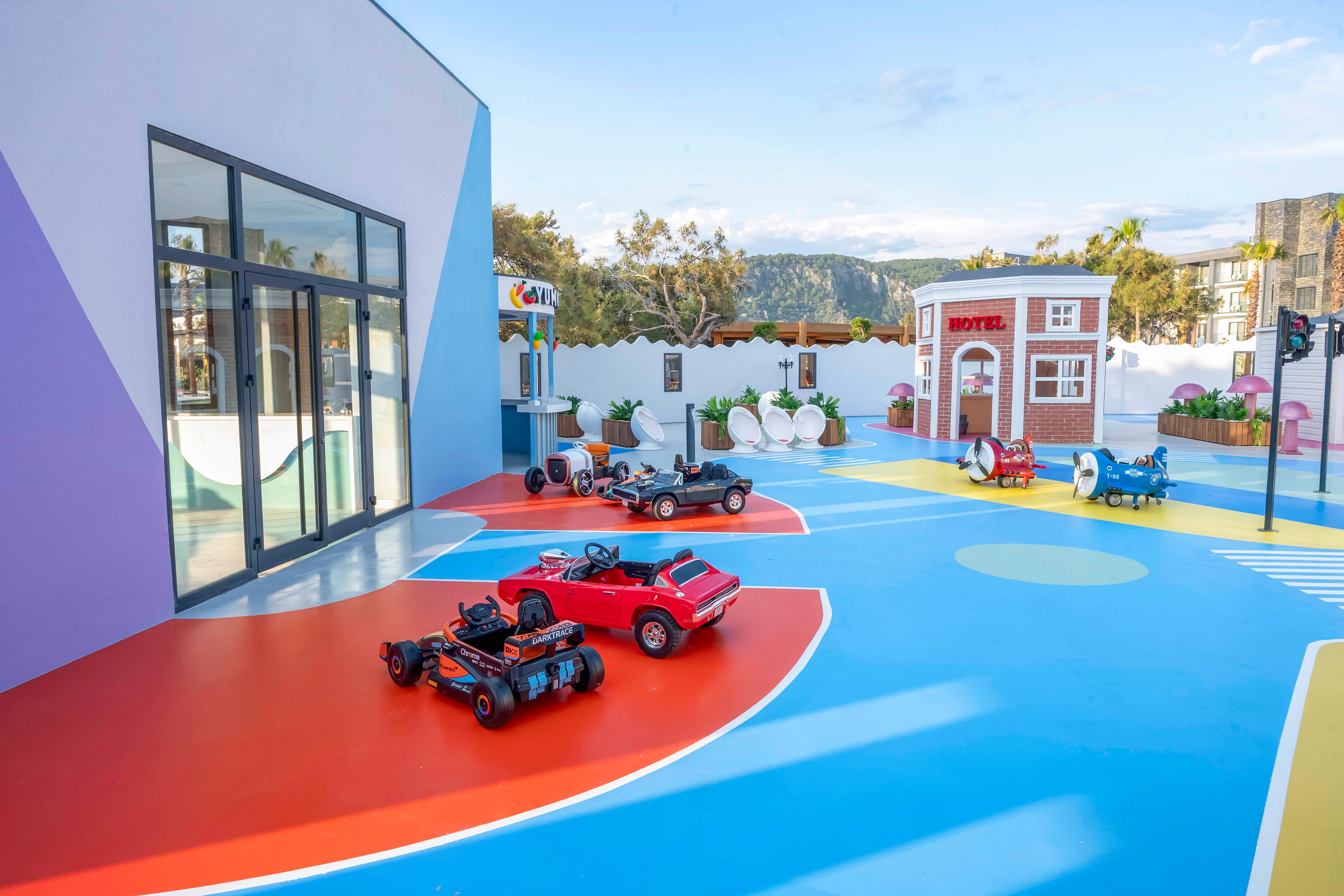 A vibrant outdoor play area features several small ride-on toy cars arranged on a brightly painted surface. The ground is divided into bold sections of blue, red, orange, and purple, creating a playful racetrack-like environment. In the background, a small playhouse styled as a motel and other whimsical structures add to the imaginative setting. The scene is sunny and open, suggesting a family-friendly recreational space for children.