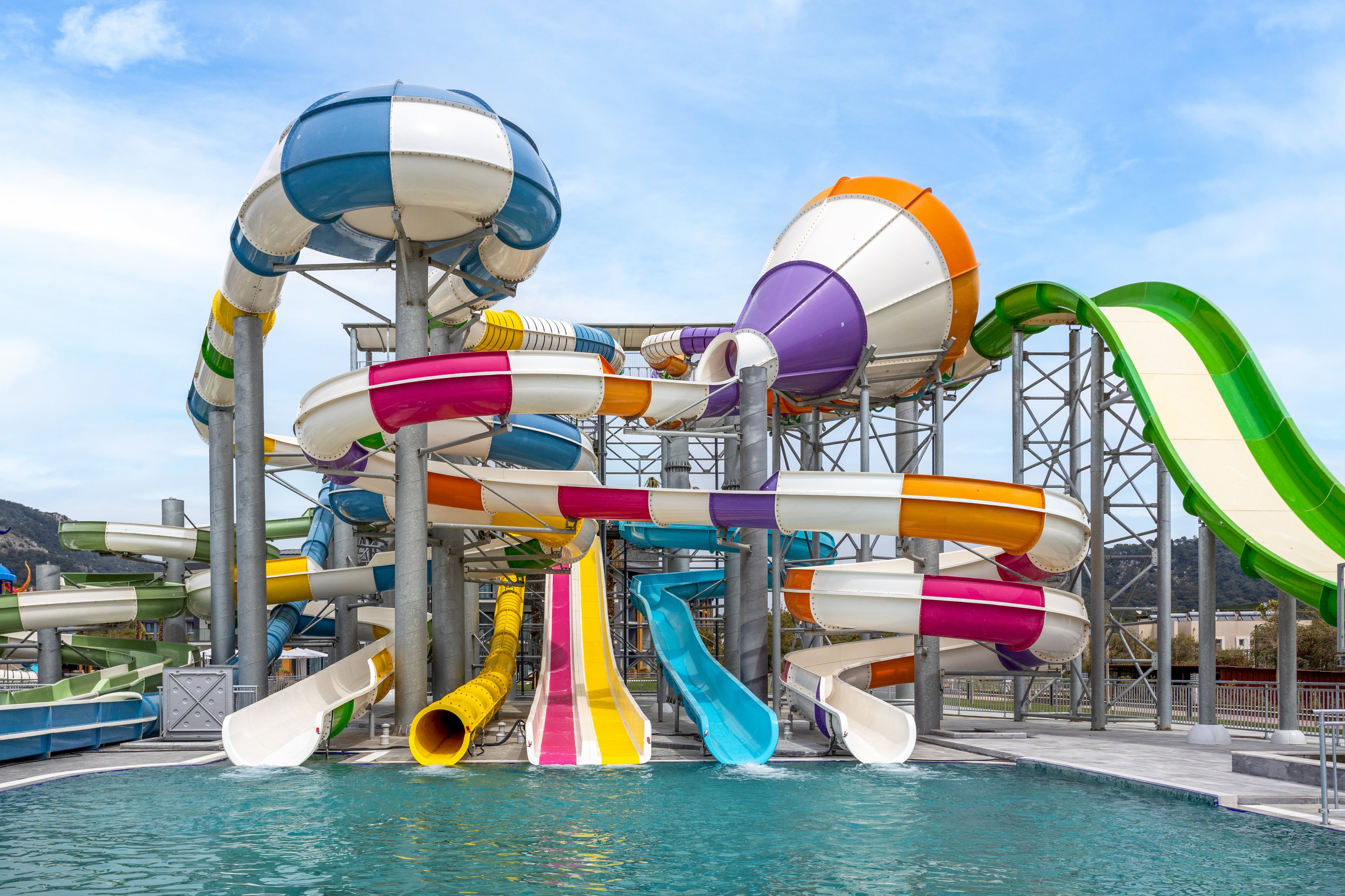 A large complex of colorful water slides towers above a clear blue swimming pool. Multiple twisting tubes and open slides in bright stripes of yellow, orange, purple, green, and blue create a dynamic, playful structure. The scene is set outdoors under a blue sky with light clouds, with metal supports and distant hills visible in the background. The overall atmosphere suggests a fun, energetic day at a modern water park attraction.