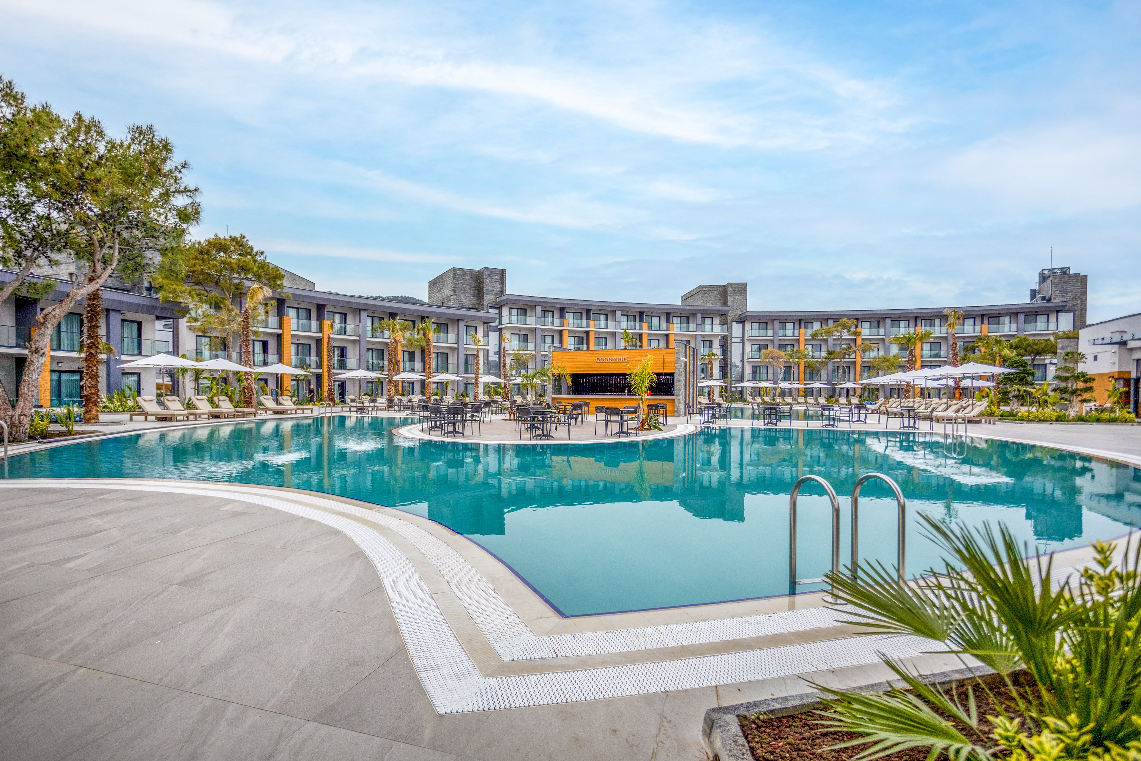 A contemporary resort hotel features a large circular outdoor swimming pool as its central focal point. The multi-story building curves around the pool, with balconies overlooking the water and poolside loungers shaded by umbrellas. Clear blue water reflects the surrounding architecture and sky, while stainless steel ladders provide access to the pool. Lush greenery and palm plants add a tropical touch to the clean, modern design.