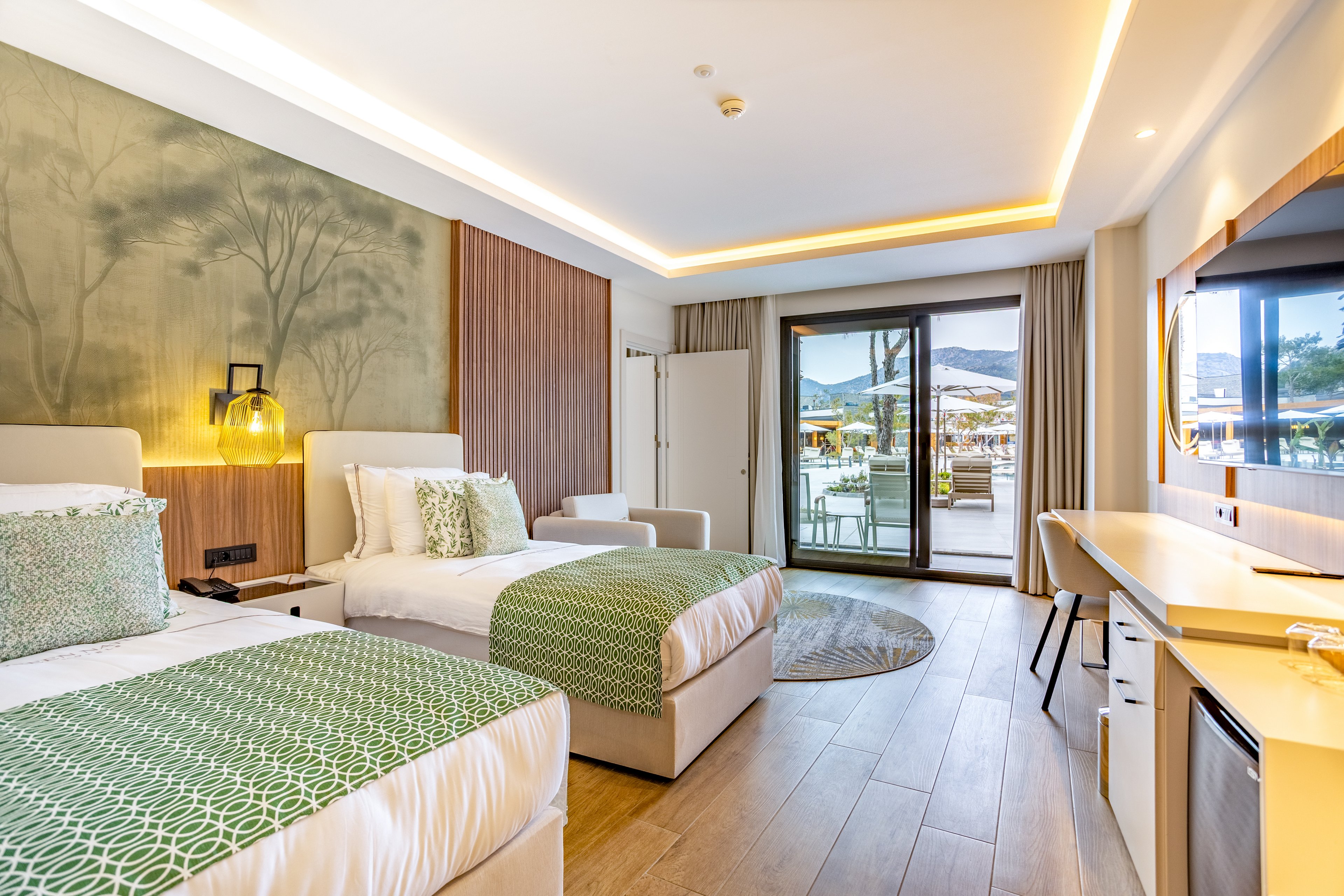 Contemporary hotel room featuring two neatly made twin beds with green patterned throws and accent pillows. The space includes a long built-in desk, mini fridge, and warm wood flooring. Large sliding glass doors open to a balcony with outdoor seating and a view of mountains and a pool area. Soft recessed lighting and a nature-inspired wall mural create a calm, inviting atmosphere.