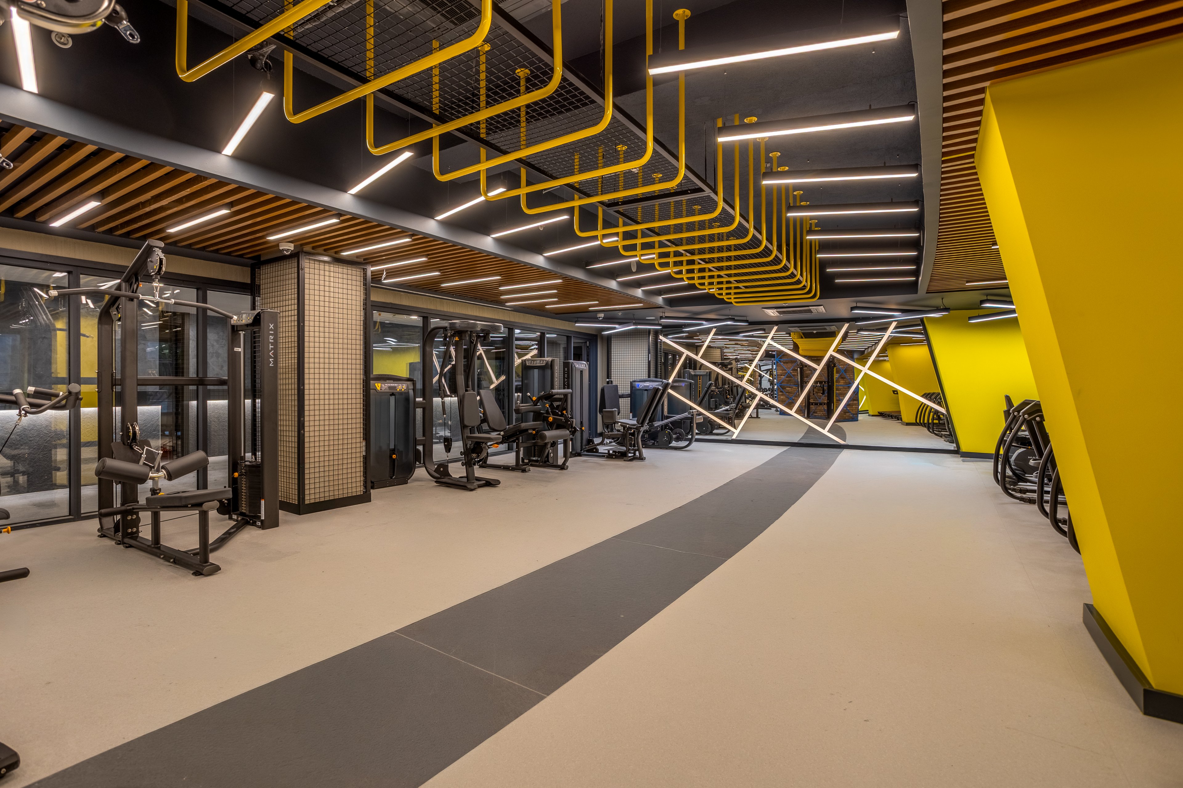 A contemporary indoor gym features various strength and cardio machines arranged along a spacious walkway. The room has bold yellow accent walls, geometric ceiling elements, and linear LED lighting. Large windows and mirrors enhance the sense of space and brightness. The overall design feels clean, organized, and ready for workouts in a modern fitness facility.