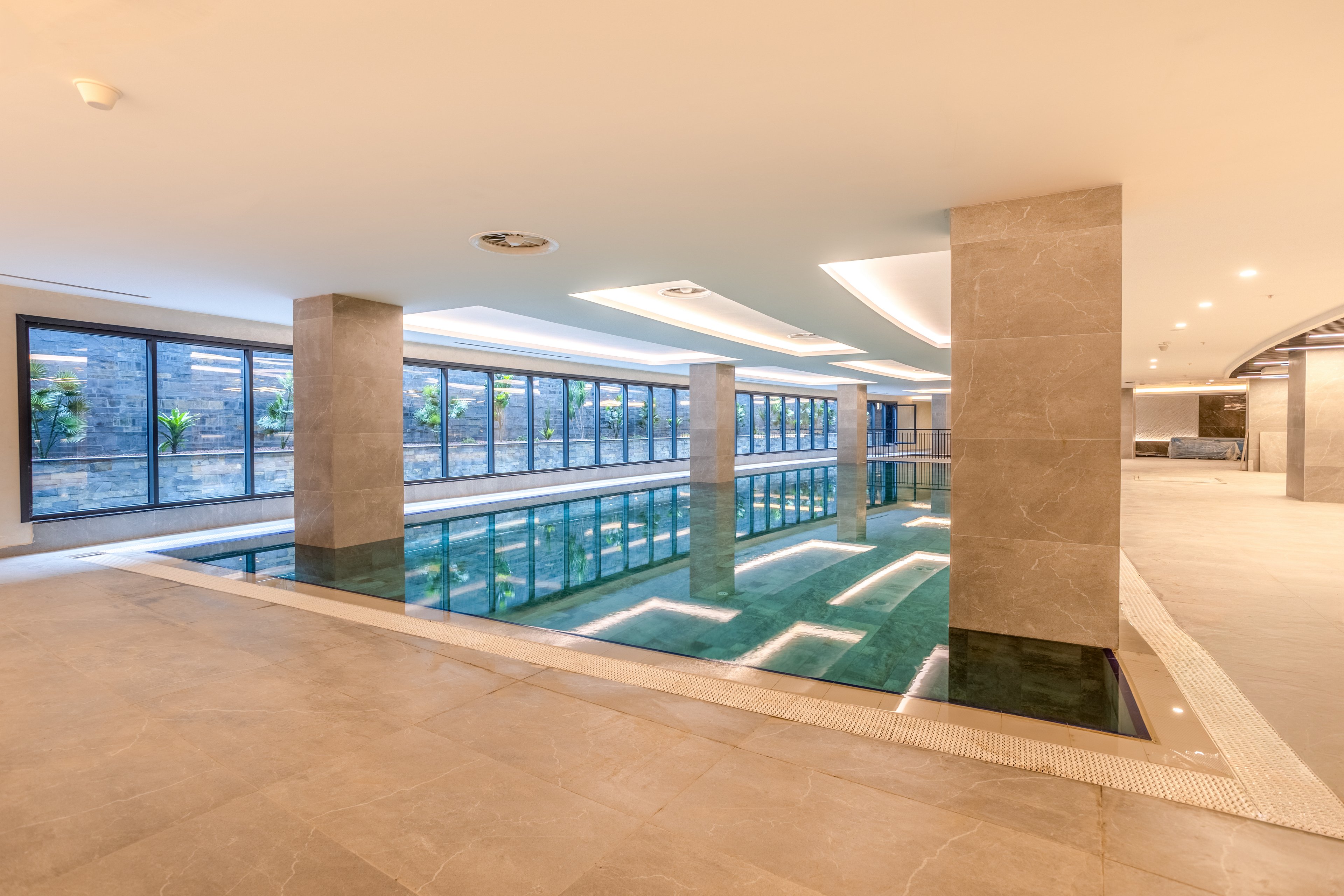 A sleek indoor swimming pool is set within a contemporary building interior. Large floor-to-ceiling windows line one side, allowing natural light to reflect off the calm water. Neutral-toned tiles and recessed ceiling lights create a clean, minimalist aesthetic. The space appears quiet and luxurious, suitable for relaxation or exercise.