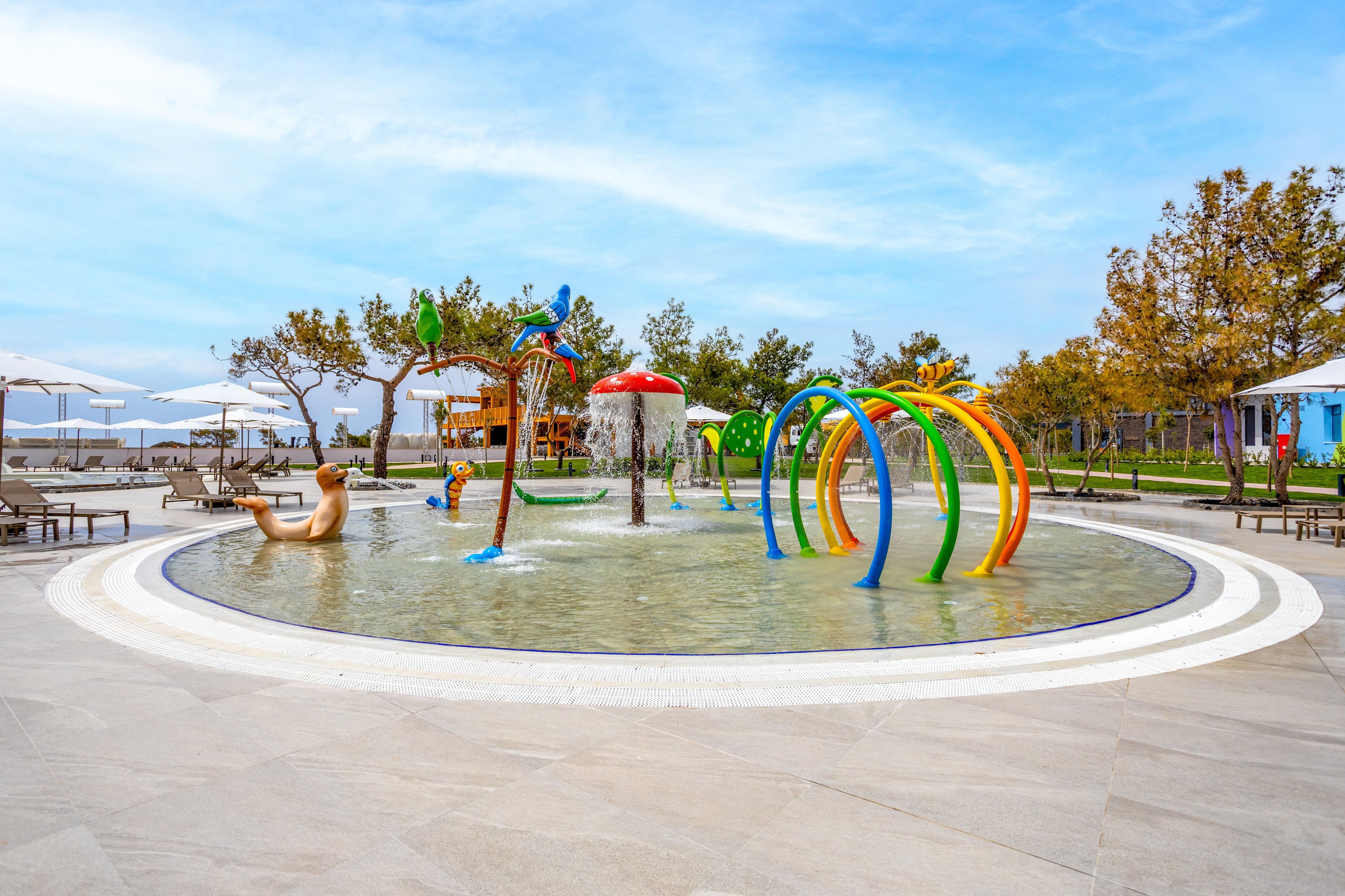 An outdoor children’s splash pad features colorful water play structures in a shallow circular pool. Arched spray rings, a red mushroom fountain, and playful animal figures create an inviting aquatic playground. Lounge chairs and umbrellas surround the area, suggesting a resort or family recreation setting. Trees and open sky in the background add to the relaxed, vacation-like atmosphere.