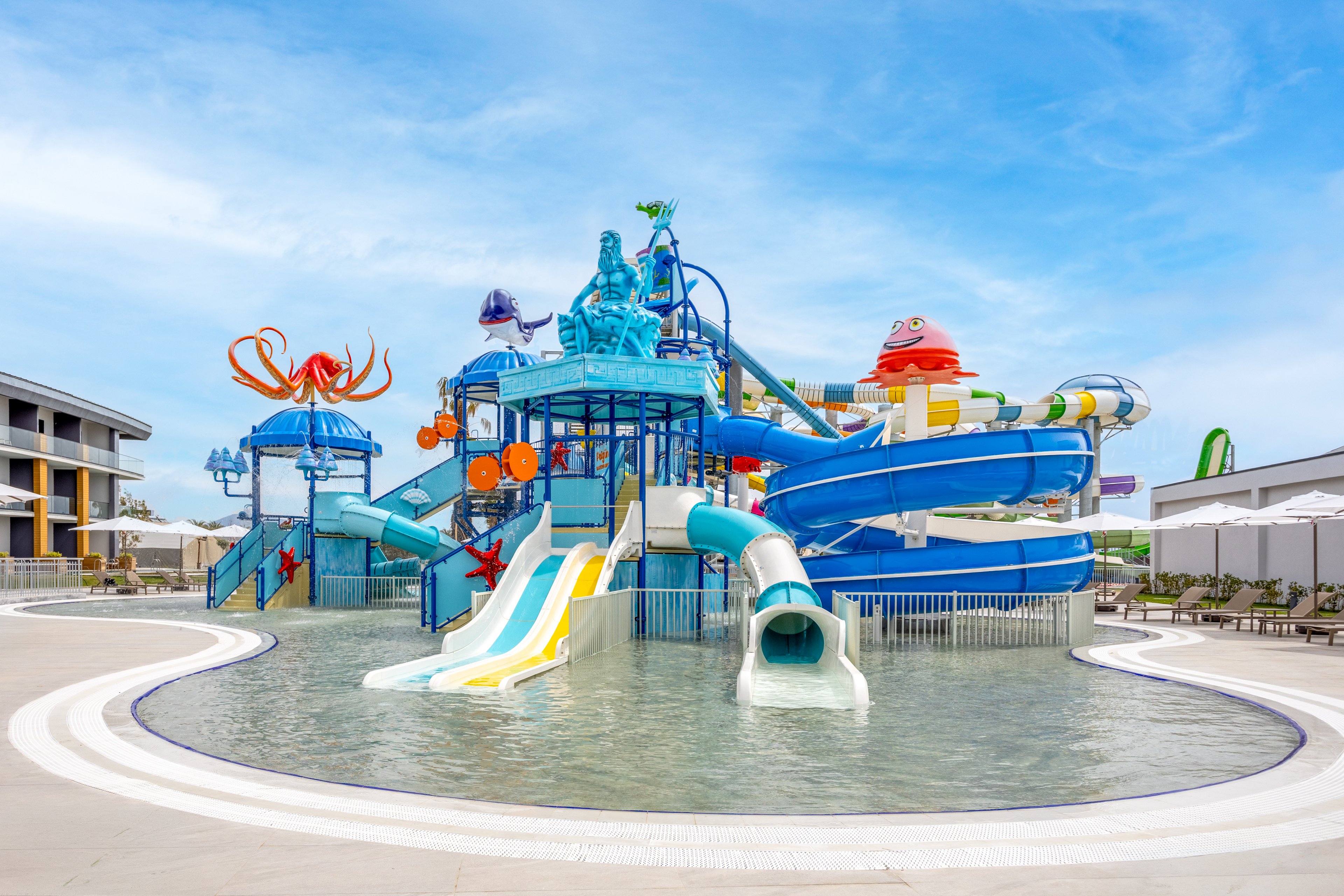 A vibrant water playground features multiple twisting slides and interactive structures in a shallow pool. Bright blue, orange, and yellow elements create a playful aquatic theme with cartoon sea creatures and water sprayers. The attraction is set in an outdoor resort area with modern buildings and lounge chairs in the background. Calm water and clear skies suggest a family-friendly vacation environment.