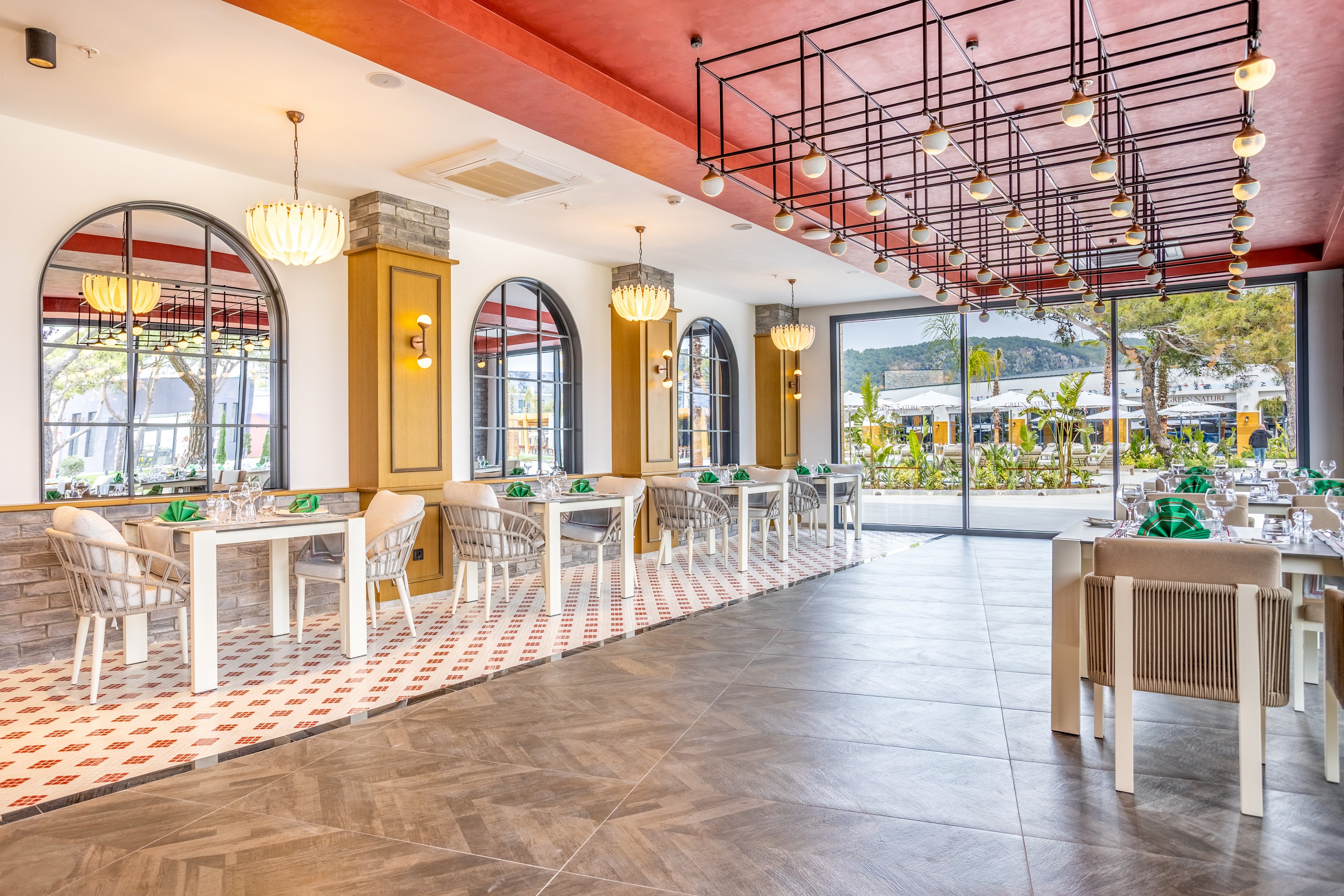 A contemporary restaurant dining room features neatly arranged tables and wicker chairs. Large arched mirrors and pendant chandeliers line one wall, while floor-to-ceiling windows open to a sunny outdoor terrace and garden. The space combines patterned tile and wood-look flooring with a bold red ceiling and geometric light fixtures. Natural light floods the room, creating an inviting and airy atmosphere ready for guests.