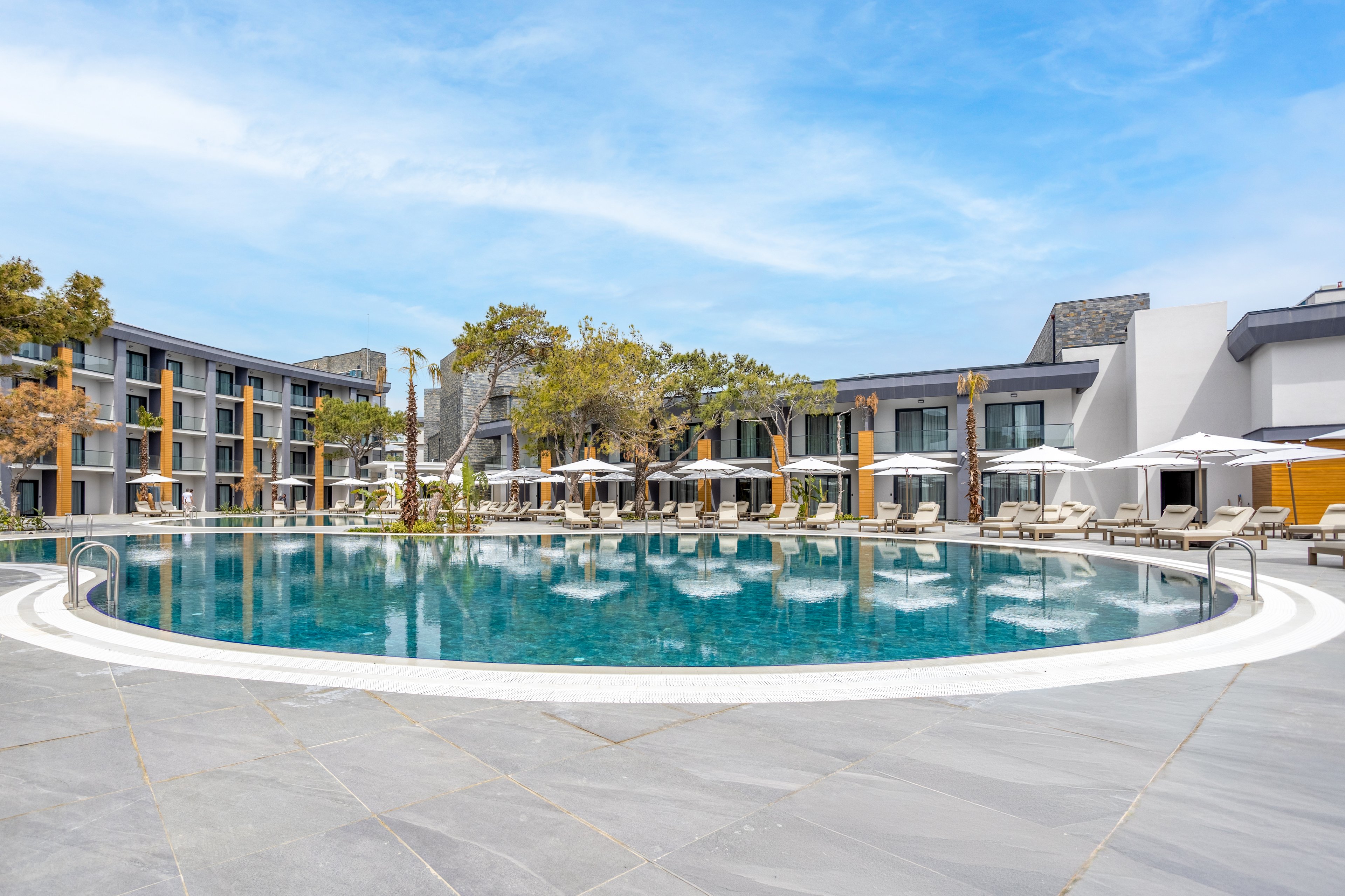 A large circular swimming pool sits at the center of a contemporary resort hotel. Sun loungers and white umbrellas line the poolside, creating a relaxed vacation atmosphere. Low-rise modern buildings with balconies surround the pool area, framed by a few trees. The bright blue sky and calm water emphasize a clean, tranquil, and inviting setting.