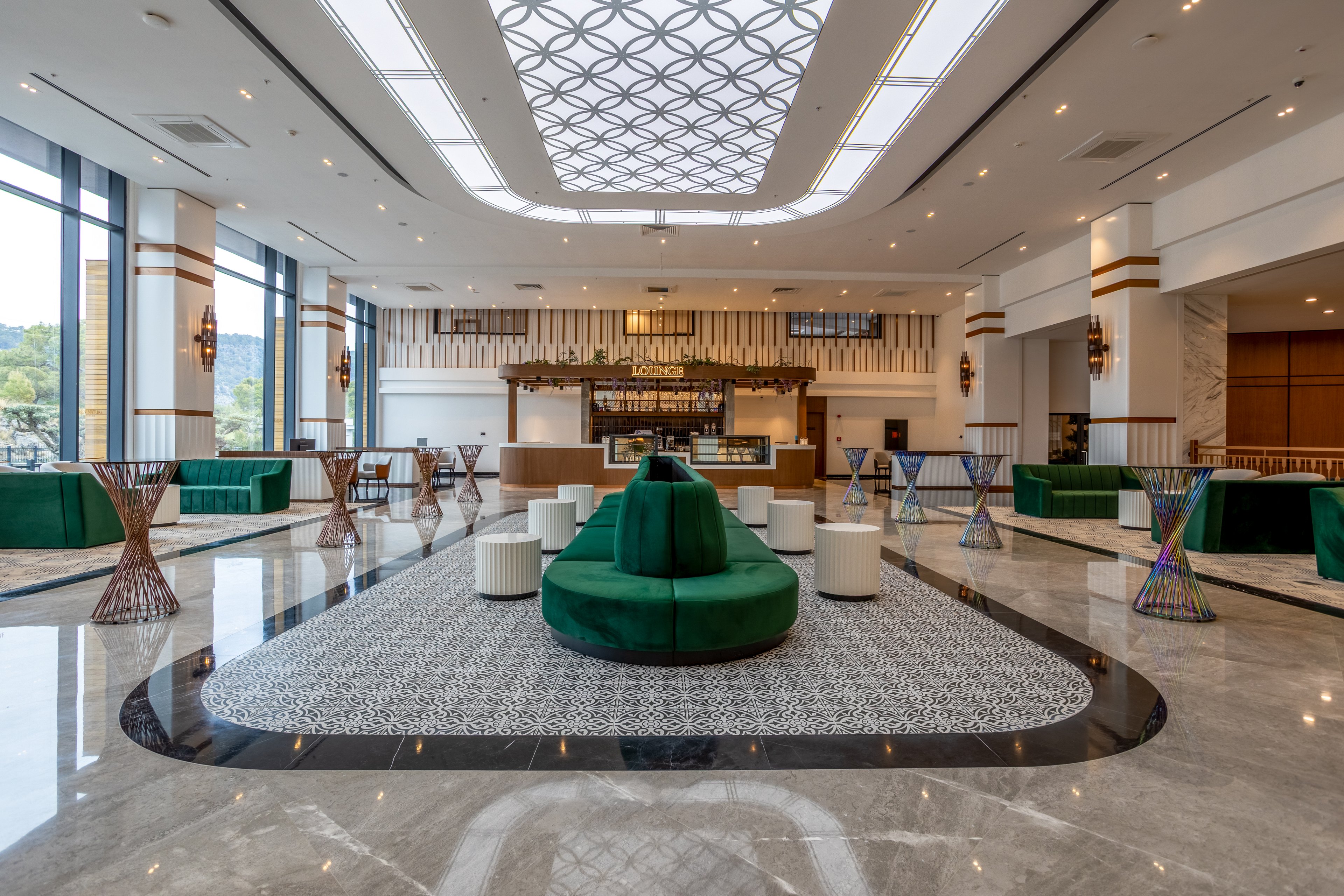 A spacious, contemporary hotel lobby features a central circular green sofa surrounded by small white stools. The area is framed by polished marble floors and a patterned rug, leading toward a stylish bar at the back. Large floor-to-ceiling windows on the left bring in natural light and views of greenery outside. Decorative side tables and geometric ceiling lighting add a sophisticated, upscale atmosphere.