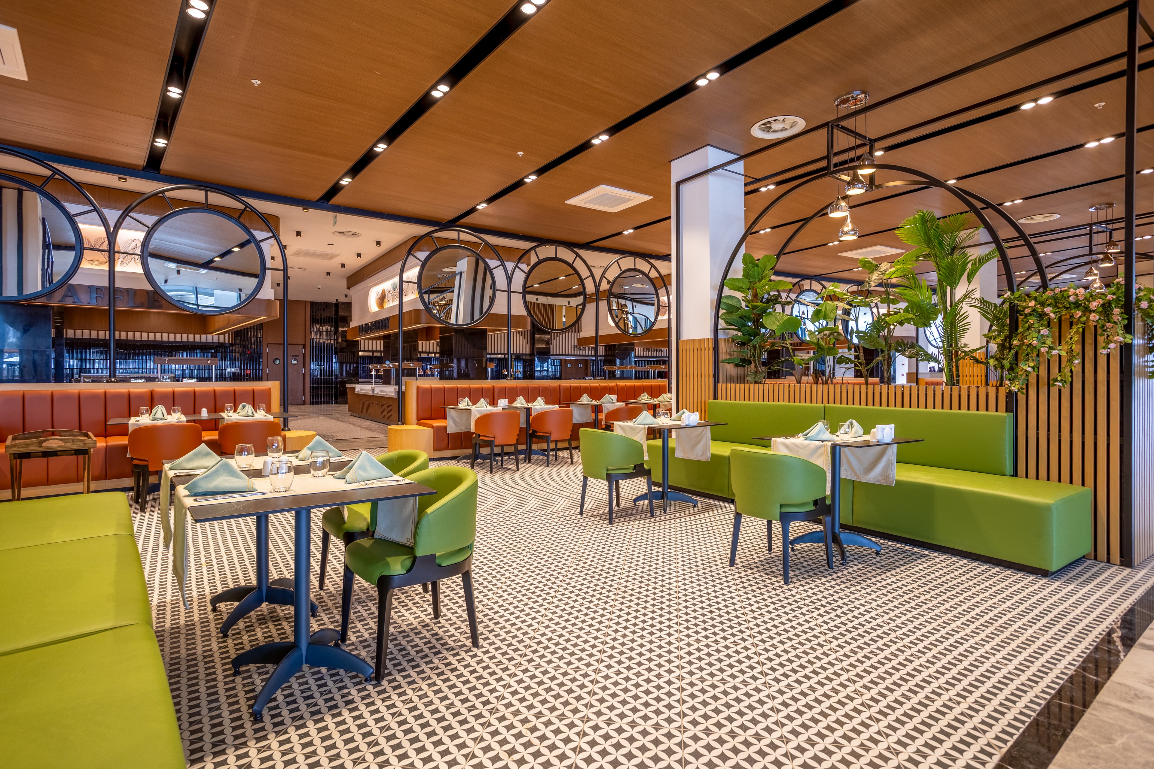 A contemporary restaurant dining area features neatly arranged tables with folded napkins and glassware. Bright green chairs and bench seating contrast with warm wood ceilings and orange banquettes. Large circular mirrors and black metal frames divide the space, while potted plants add a touch of greenery. The patterned tile floor and soft ambient lighting create a stylish yet welcoming atmosphere.