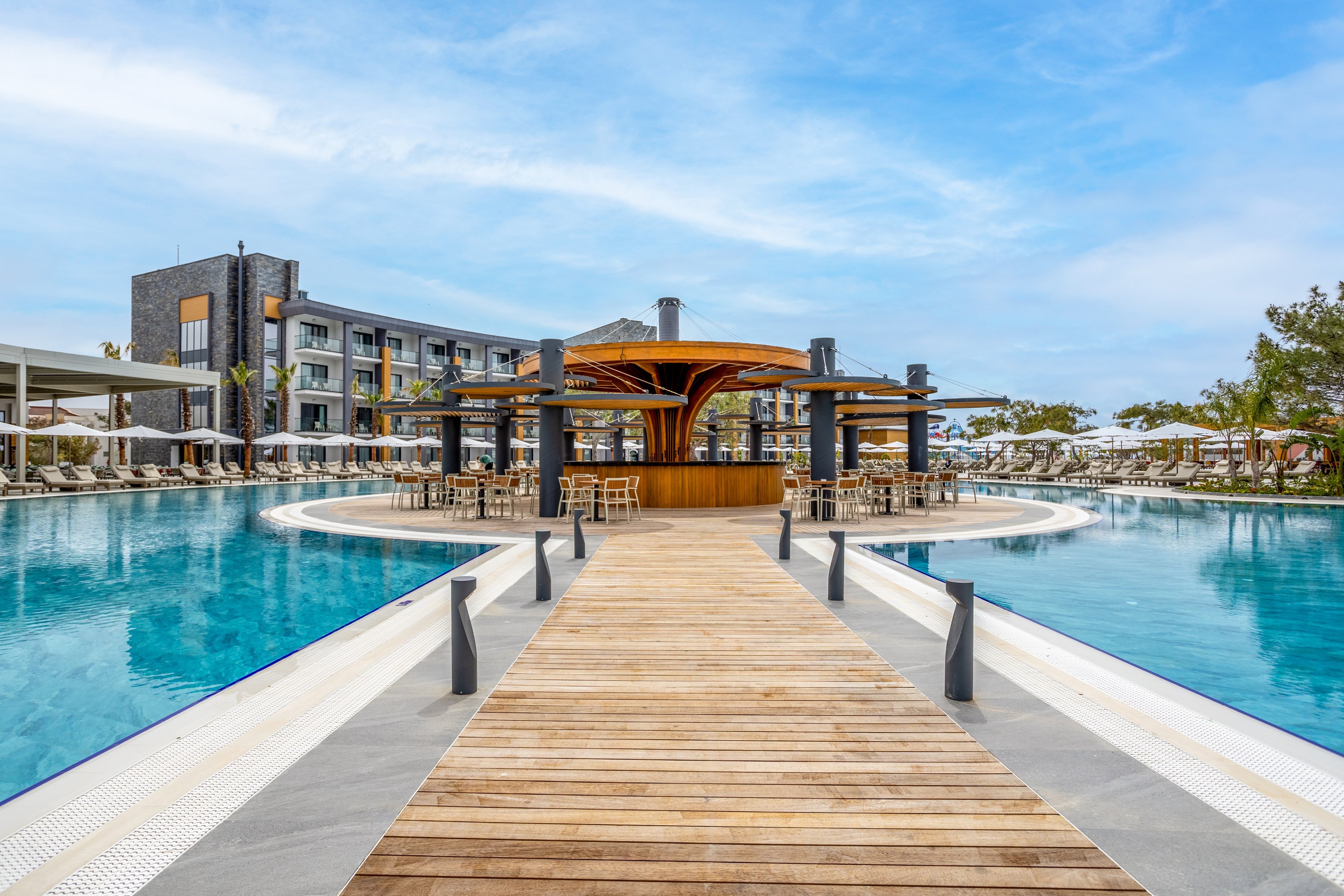 A large resort swimming pool features a wooden walkway leading to a circular island bar at its center. Modern hotel buildings and rows of sun loungers with umbrellas surround the pool, creating a relaxing vacation atmosphere. The clear blue water reflects the sky and contemporary architecture. The scene is bright and inviting, suggesting a tropical or coastal holiday destination.