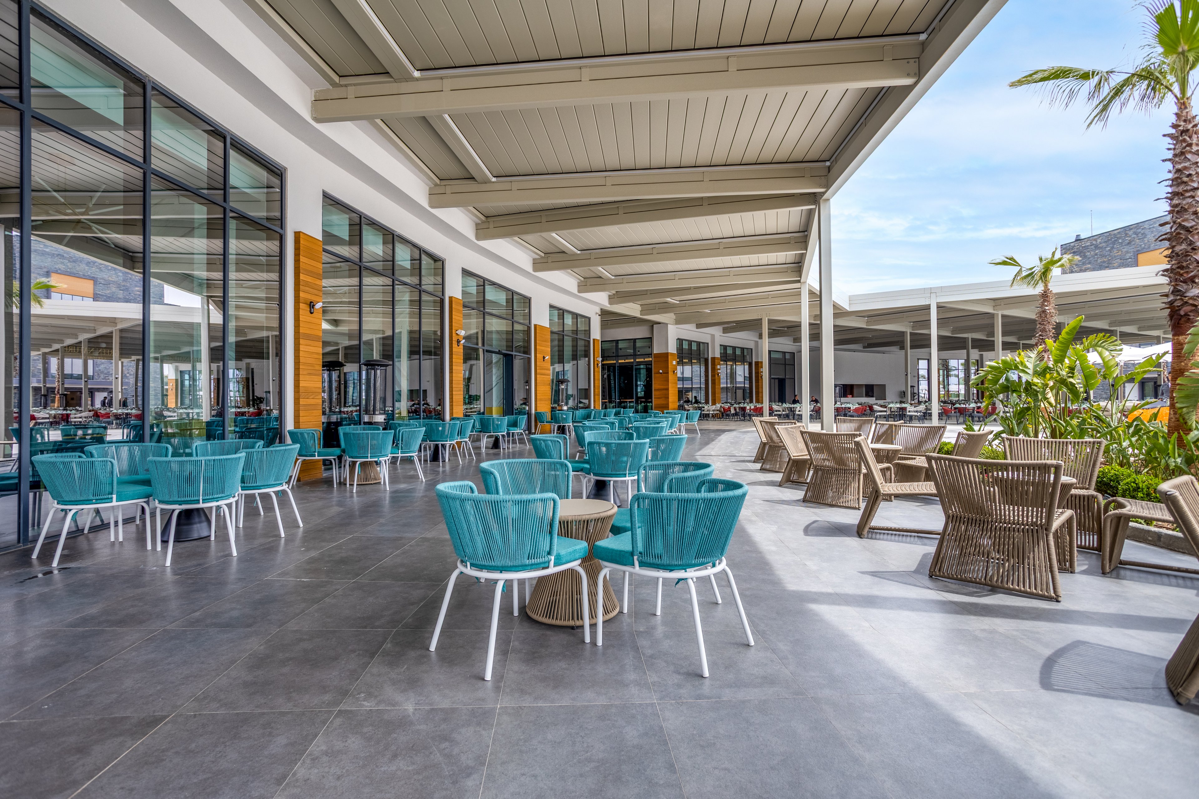 A spacious outdoor terrace features numerous turquoise chairs and white tables arranged neatly under a large covered patio. Floor-to-ceiling windows line the building, reflecting the bright sky and surrounding structures. The area is paved with smooth gray tiles and includes additional wicker-style lounge chairs near some greenery and a palm tree. The scene appears calm and inviting, suggesting a contemporary café, restaurant, or hotel relaxation area.