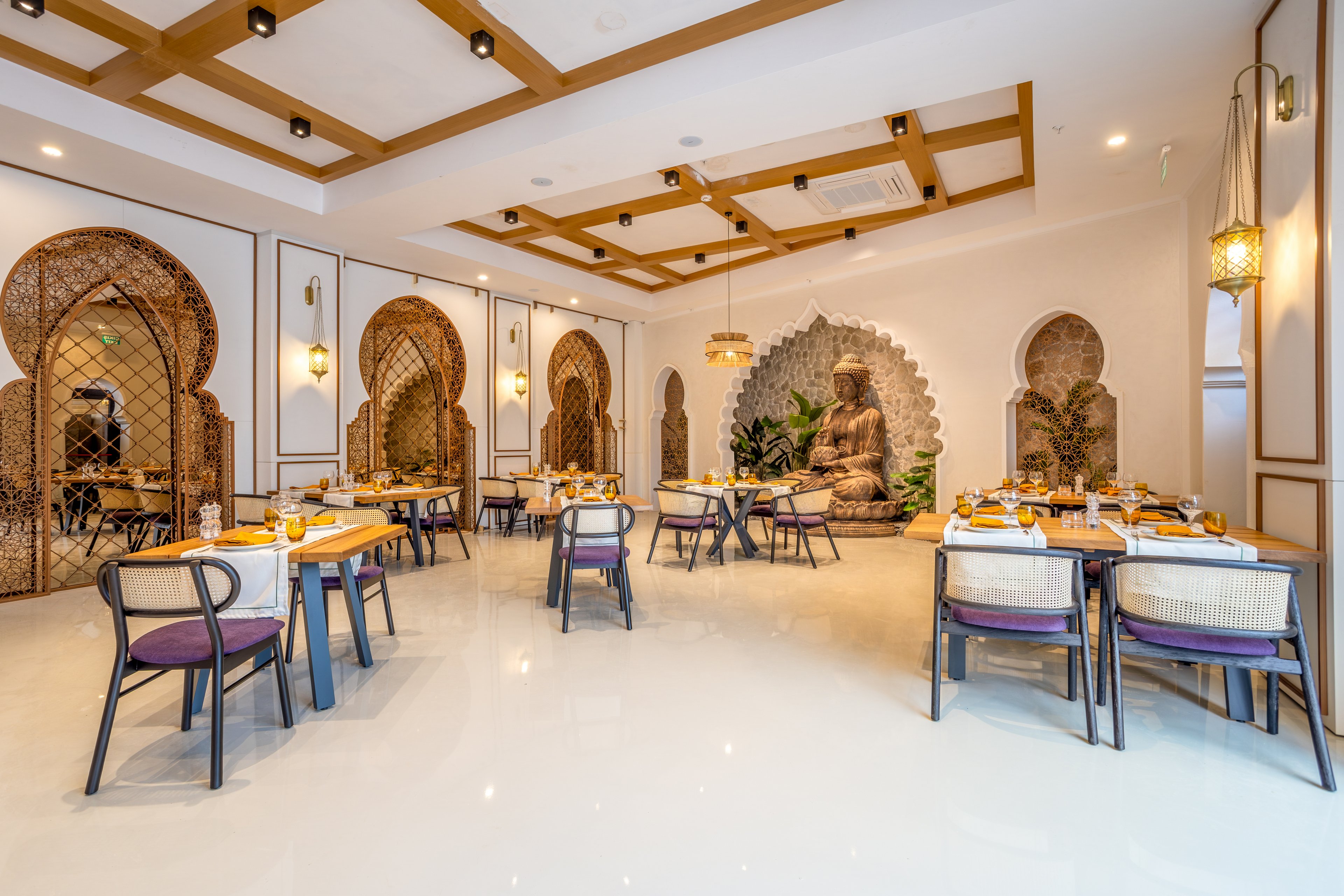 A stylish restaurant dining room features neatly arranged tables with place settings and cushioned chairs. The space has a bright, polished floor and a warm ceiling design with wooden beams and recessed lighting. Ornate arched wall niches and lattice panels add a decorative, Middle Eastern or Indian-inspired touch. A large Buddha statue surrounded by plants serves as a focal point, creating a serene and inviting atmosphere.