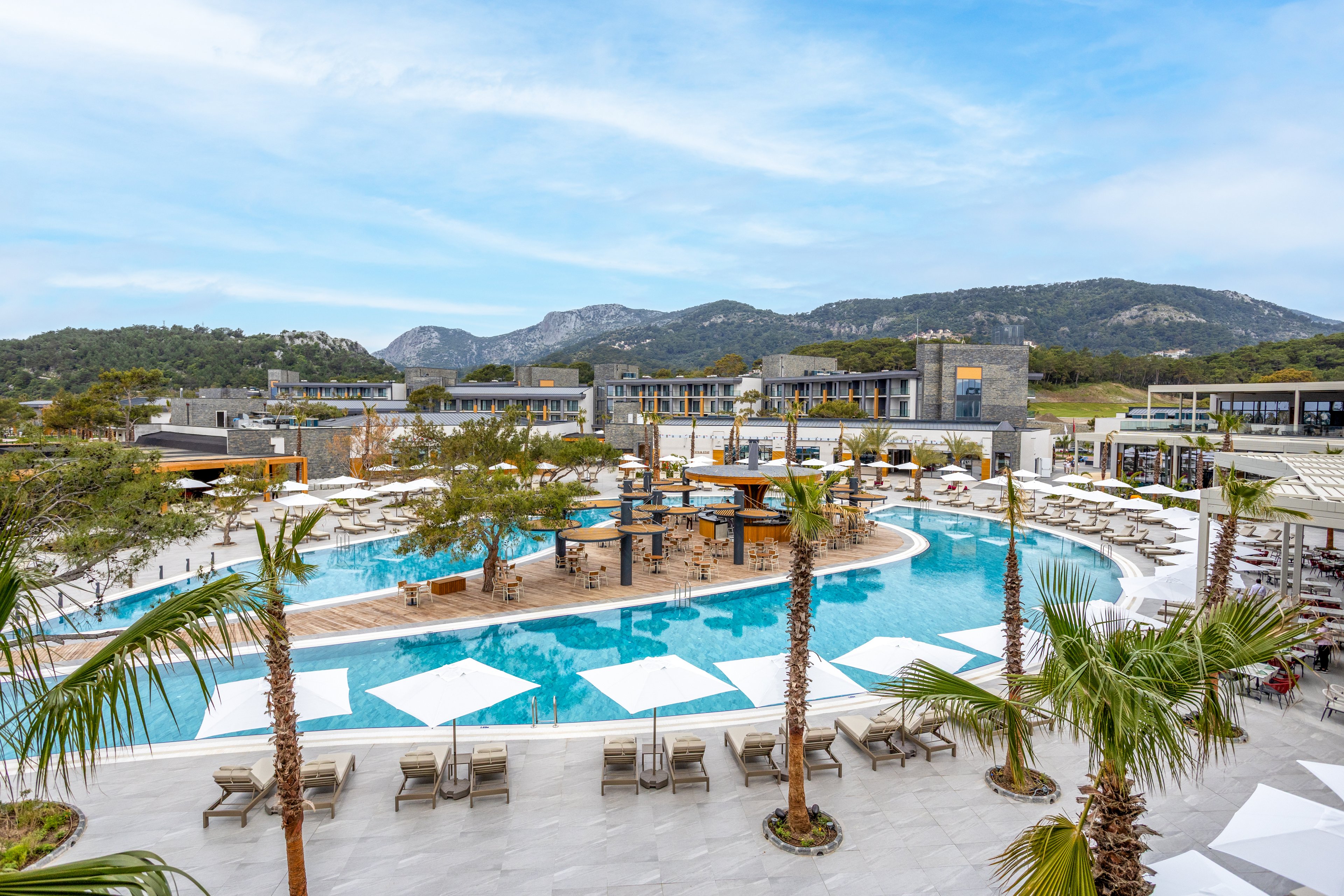 A large, contemporary resort pool dominates the scene, surrounded by sun loungers and umbrellas. Palm trees and small islands break up the water, adding a tropical feel to the modern architecture. Low-rise hotel buildings frame the pool area, with hills and greenery visible in the background. The bright daylight and blue sky create a relaxed vacation atmosphere.