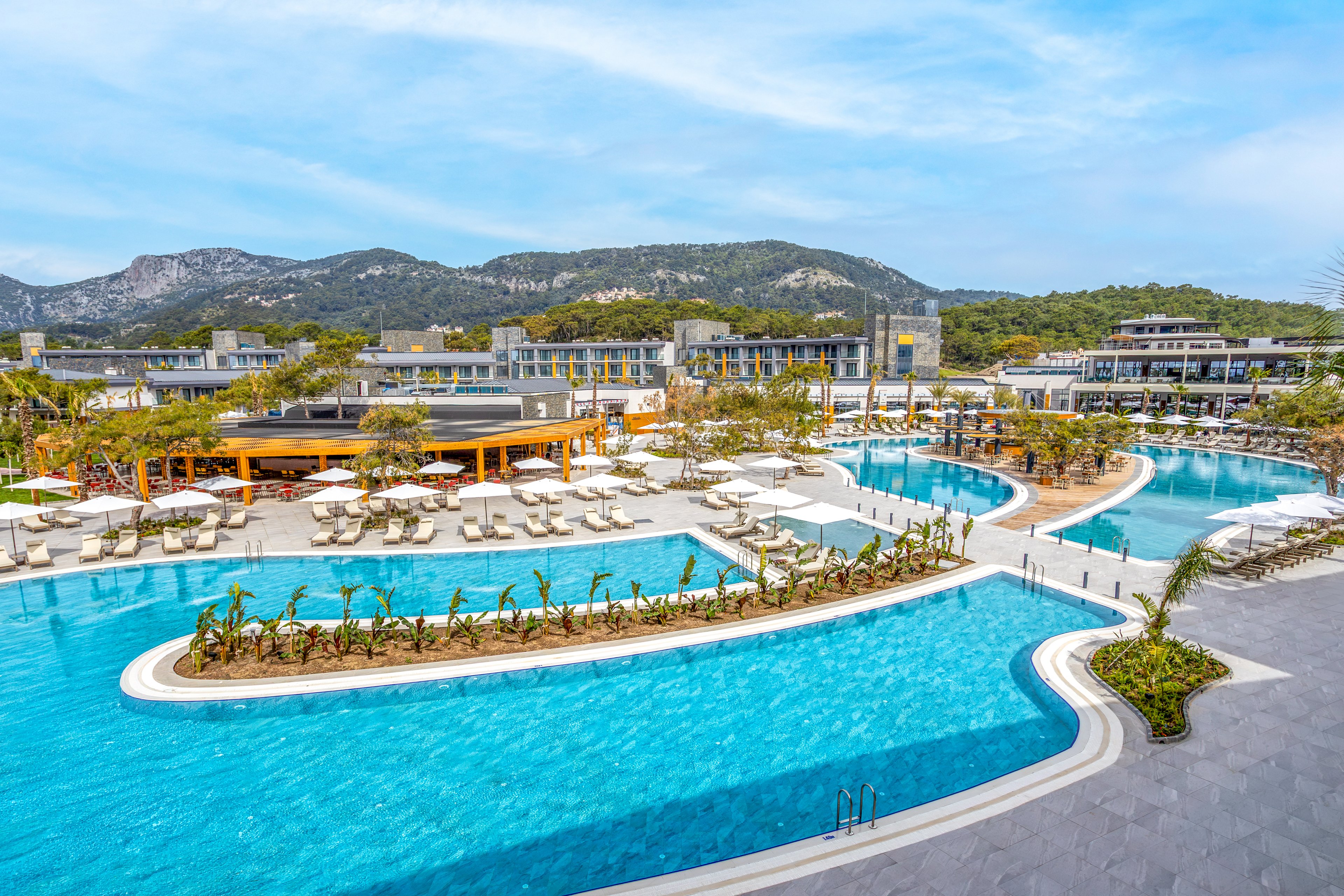 A contemporary resort features a large, winding outdoor swimming pool surrounded by sun loungers and umbrellas. Low-rise hotel buildings frame the pool area, set against a backdrop of green hills and mountains. The bright blue water contrasts with the neutral tones of the terraces and pathways, creating a clean, modern aesthetic. The scene suggests a tranquil vacation destination focused on relaxation and leisure by the pool.