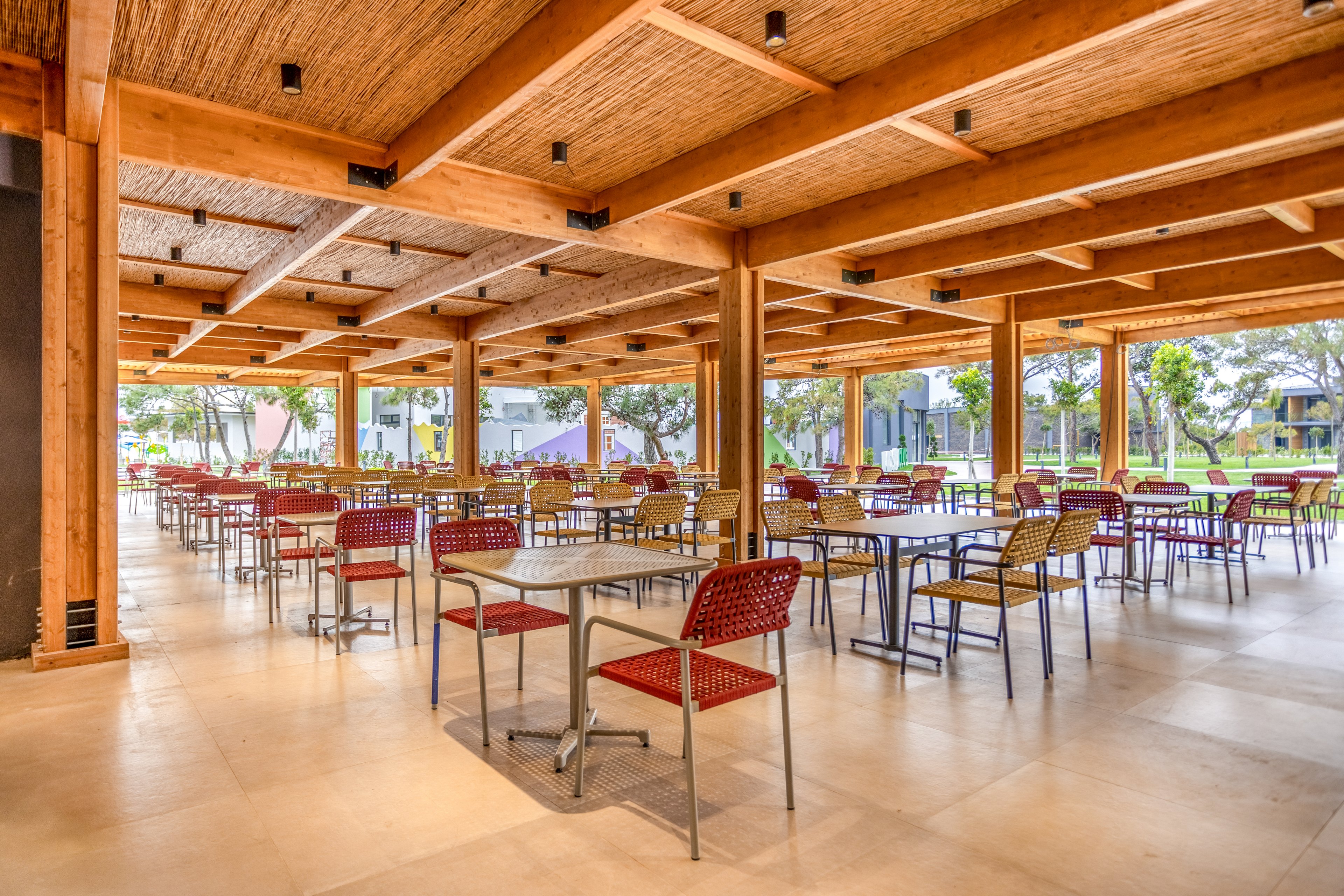 An open-air cafeteria features rows of empty tables and chairs under a wooden ceiling. Large floor-to-ceiling windows reveal a green outdoor area with trees and buildings in the background. The space is bright and airy, with natural light illuminating the neutral floor and warm wood tones. Red and wicker chairs add subtle color accents to the modern, minimalist dining area.