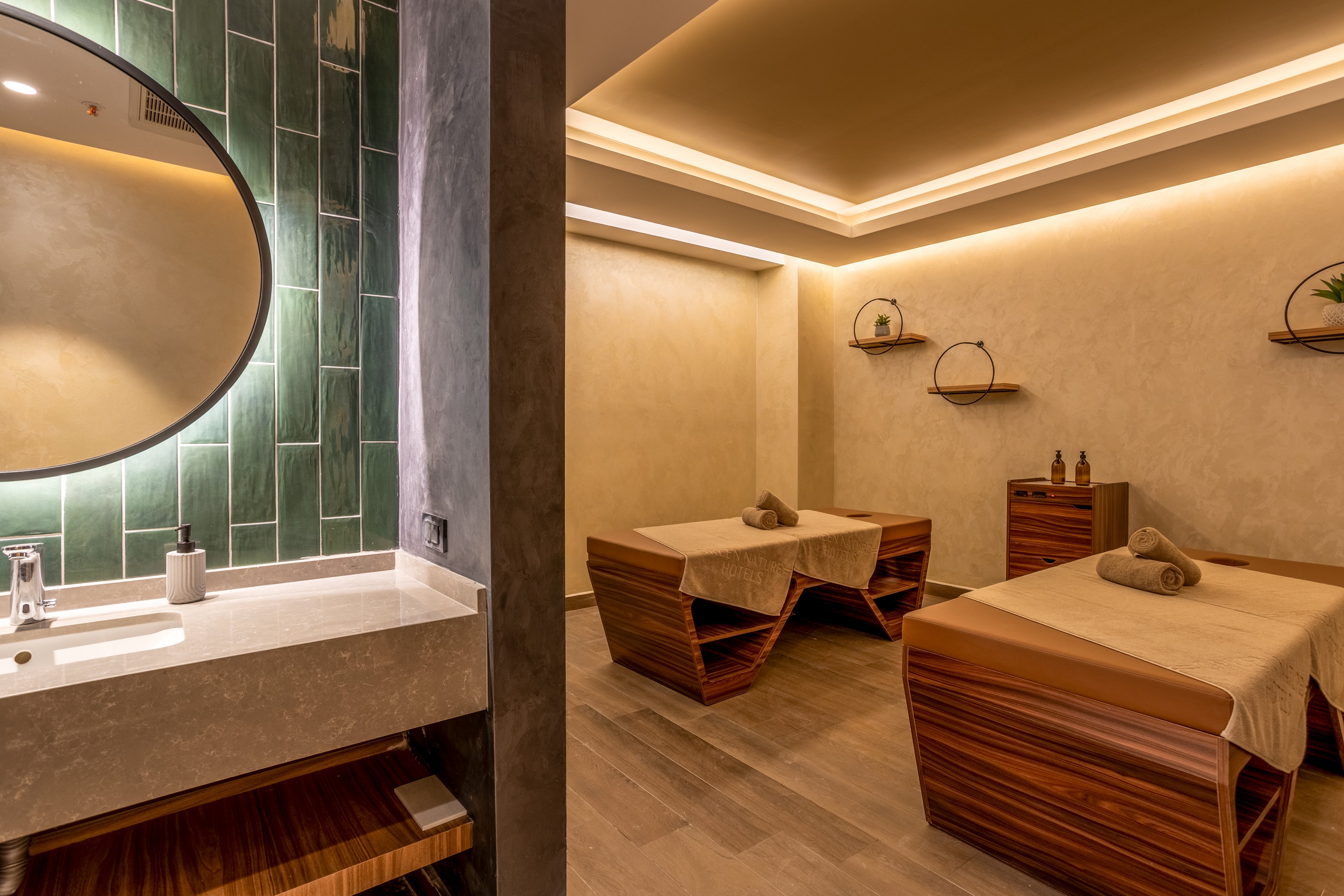 A contemporary spa treatment room features two wooden massage tables neatly prepared with beige linens and rolled towels. Warm recessed lighting and neutral-toned walls create a calm, inviting atmosphere. A vanity area with a round mirror and green tiled backsplash is visible to the left, adding a touch of color and texture. Minimal decor, including small shelves and bottles, enhances the clean, relaxing spa environment.