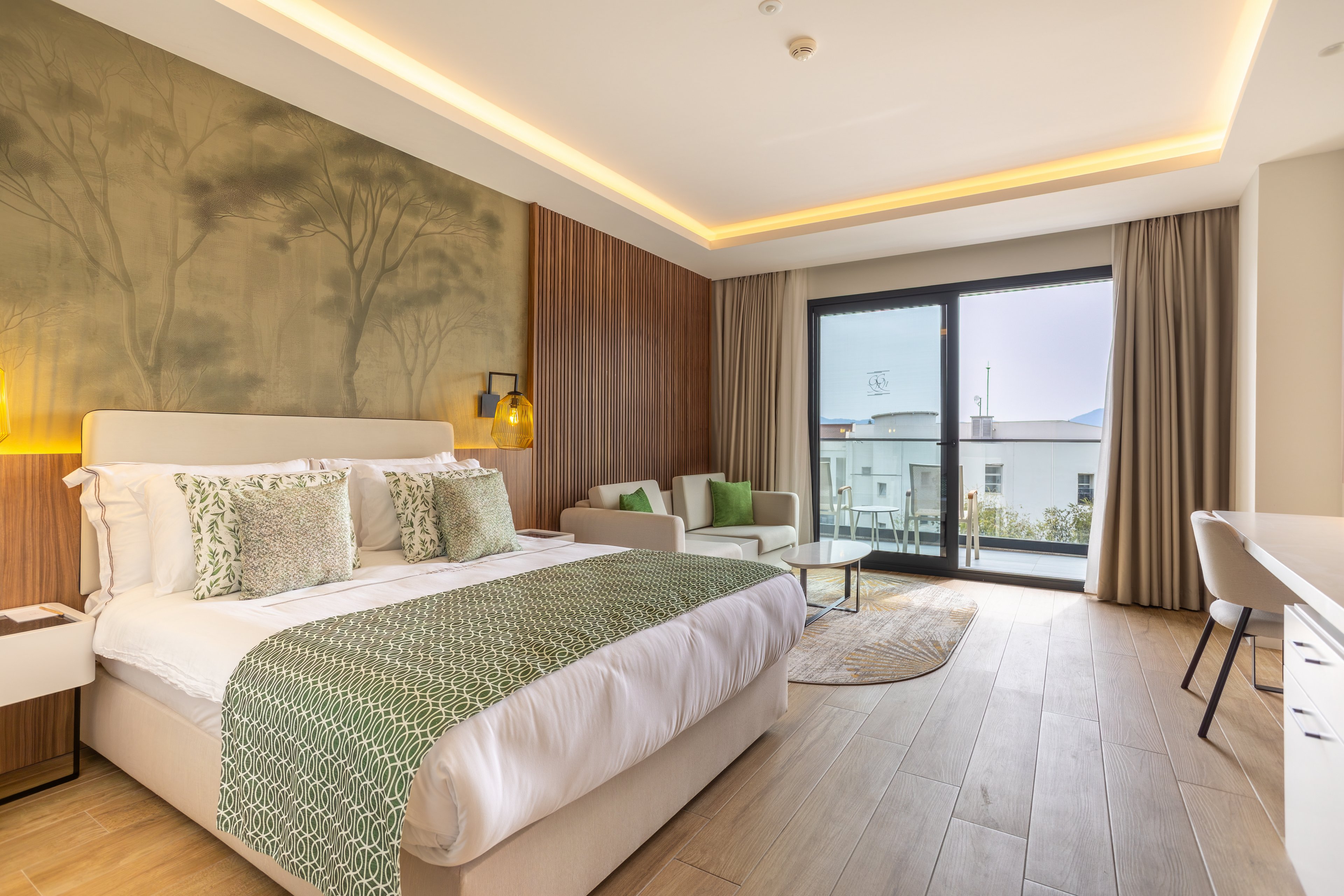 A contemporary hotel bedroom features a large neatly made bed with patterned green textiles and decorative pillows. The room includes a small seating area with a sofa, armchairs, and a round coffee table near the sliding glass doors. Warm wood paneling, soft recessed lighting, and a nature-inspired wall mural create a calm, inviting atmosphere. Outside the balcony doors, neighboring buildings and a bright sky are visible, adding natural light to the interior.