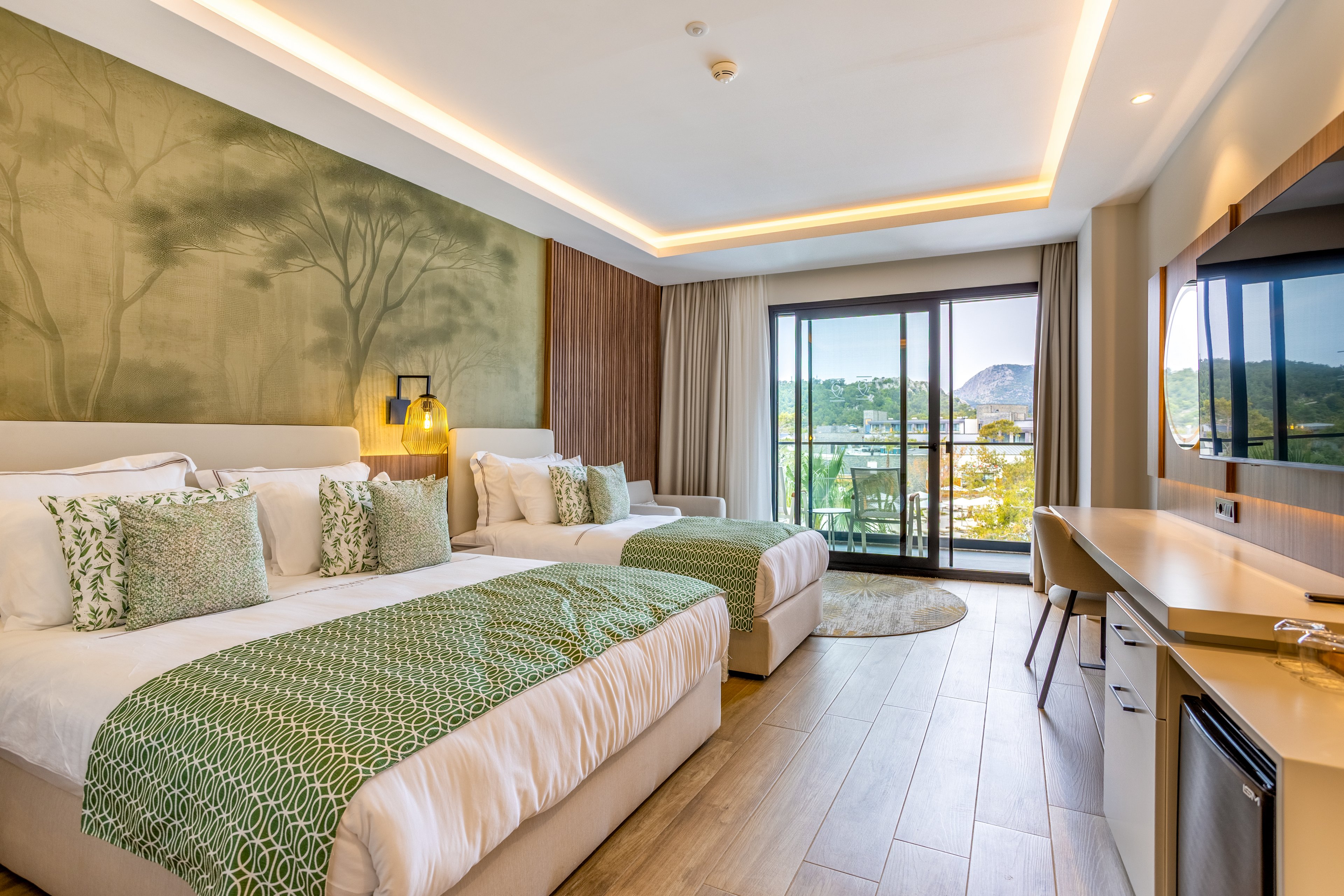 Bright contemporary hotel room featuring two large double beds with coordinated green and white bedding. The space includes a long built-in desk, flat-screen television, and a compact refrigerator. Warm recessed ceiling lighting and a nature-themed accent wall create a calm, inviting atmosphere. Floor-to-ceiling glass doors open to a balcony with views of a distant hill and surrounding landscape.