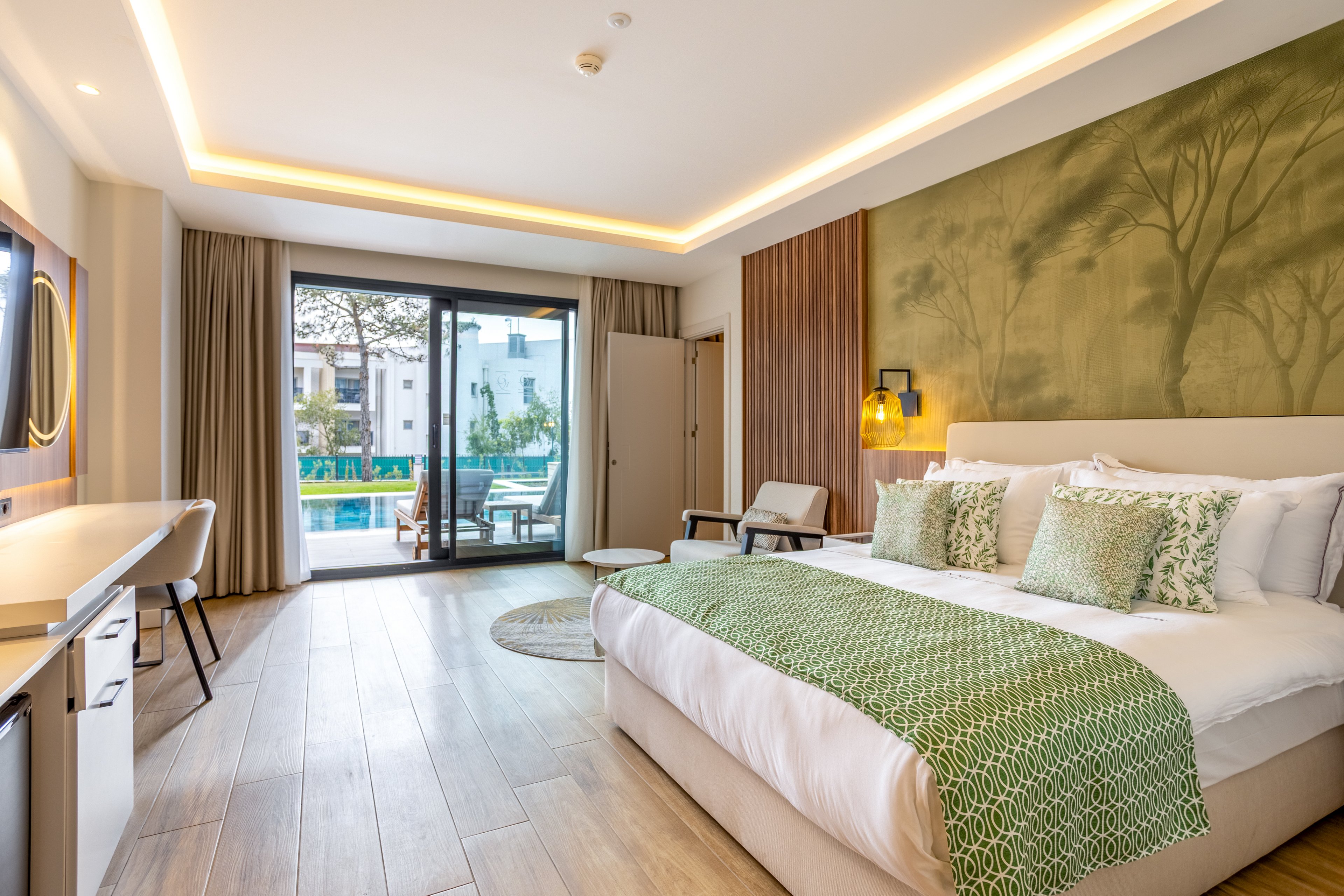 A contemporary hotel room features a large bed with green patterned bedding and multiple decorative pillows. The space includes a desk area, armchair, and warm recessed ceiling lighting that creates a cozy atmosphere. Floor-to-ceiling glass doors open onto a balcony overlooking a swimming pool and neighboring buildings. Natural tones, wood flooring, and a nature-themed wall mural contribute to a calm, resort-like setting.