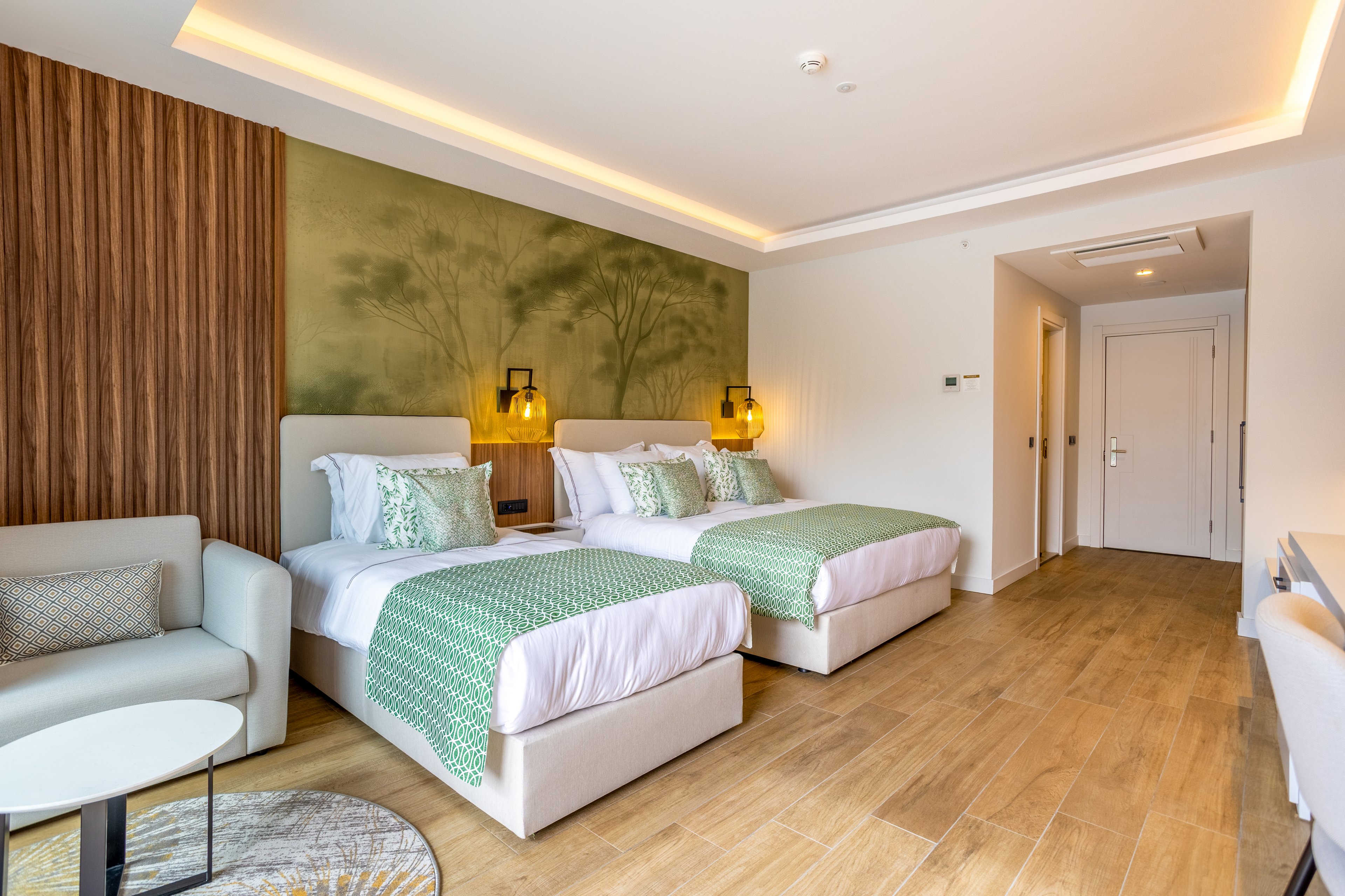 A contemporary hotel room features two double beds with white linens and green patterned throws. The space includes a light-colored sofa, round coffee table, and wooden flooring that adds warmth. A green nature-themed wall mural and wooden panel accent wall create a calm, natural ambiance. Soft recessed ceiling lighting and bedside lamps provide a cozy, inviting atmosphere, while a doorway and hallway lead to the rest of the suite.