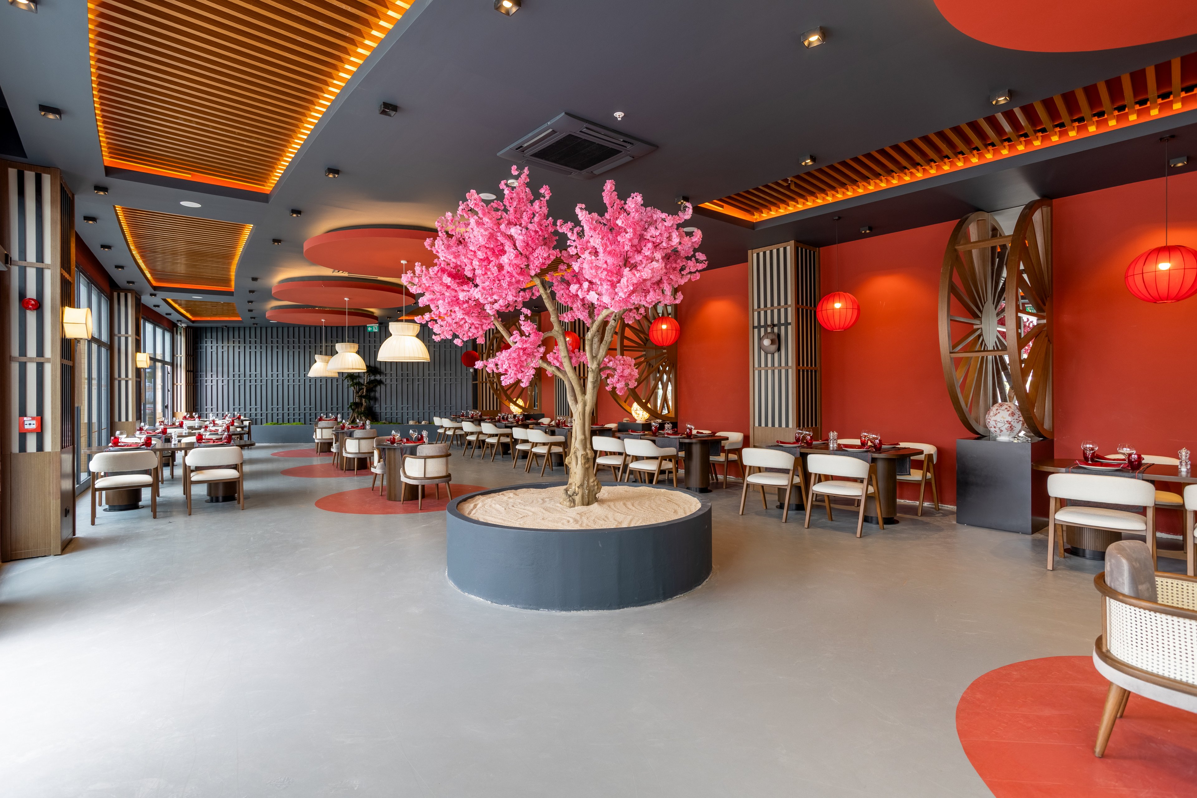 A contemporary restaurant interior features a striking artificial pink blossom tree planted in a circular planter at the center of the room. The space is decorated with red walls, wooden slatted ceiling panels, and warm pendant lighting that creates an inviting atmosphere. Dining tables and cushioned chairs are neatly arranged around the central feature, ready for guests. Red lanterns and decorative wall elements add an elegant, Asian-inspired touch to the modern design.
