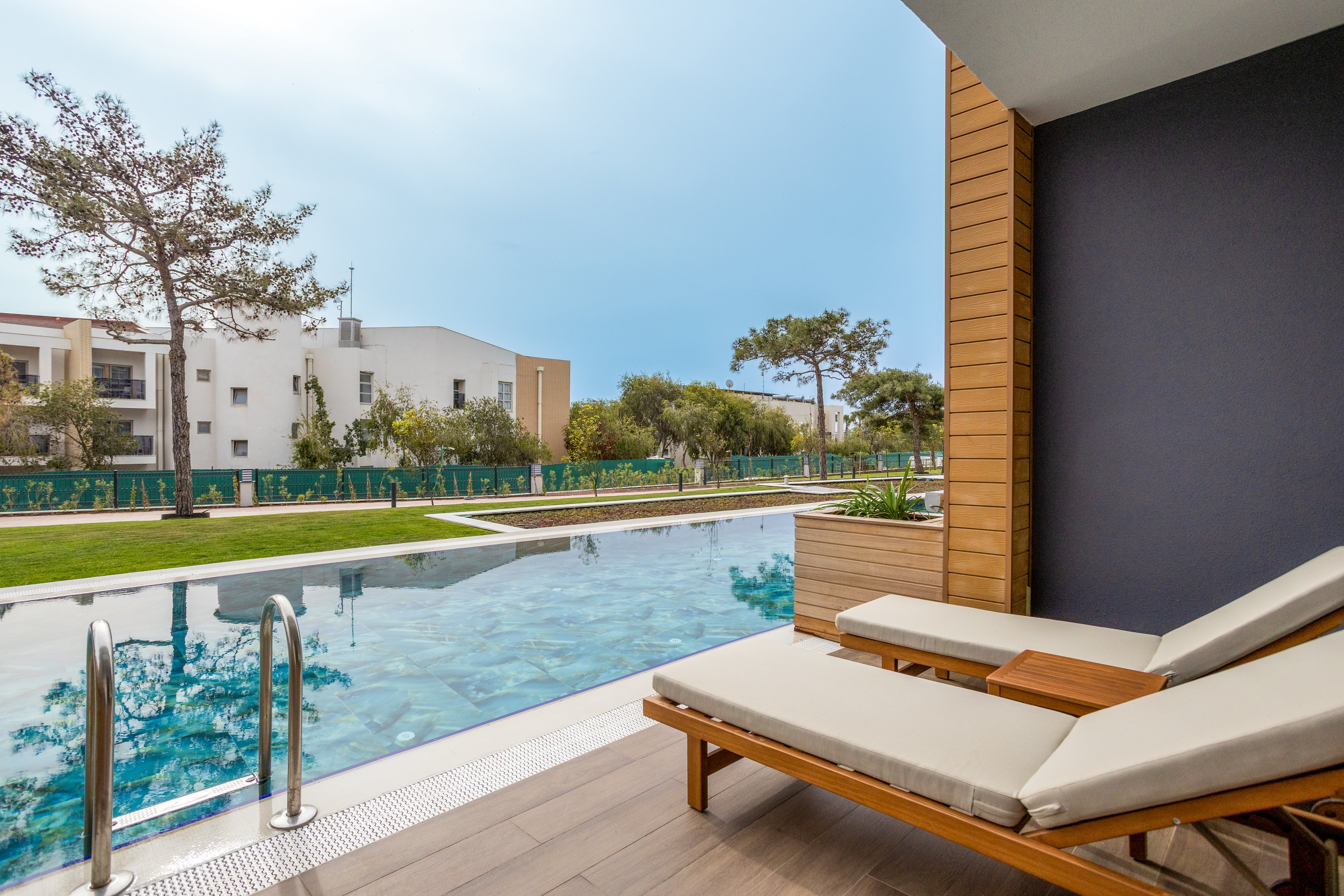 A contemporary outdoor terrace overlooks a long rectangular swimming pool. Two cushioned wooden lounge chairs sit on a wooden deck beside metal pool ladders. In the background, modern apartment buildings and green lawns with trees create a residential setting. The calm water and clear sky contribute to a relaxed, resort-like atmosphere.