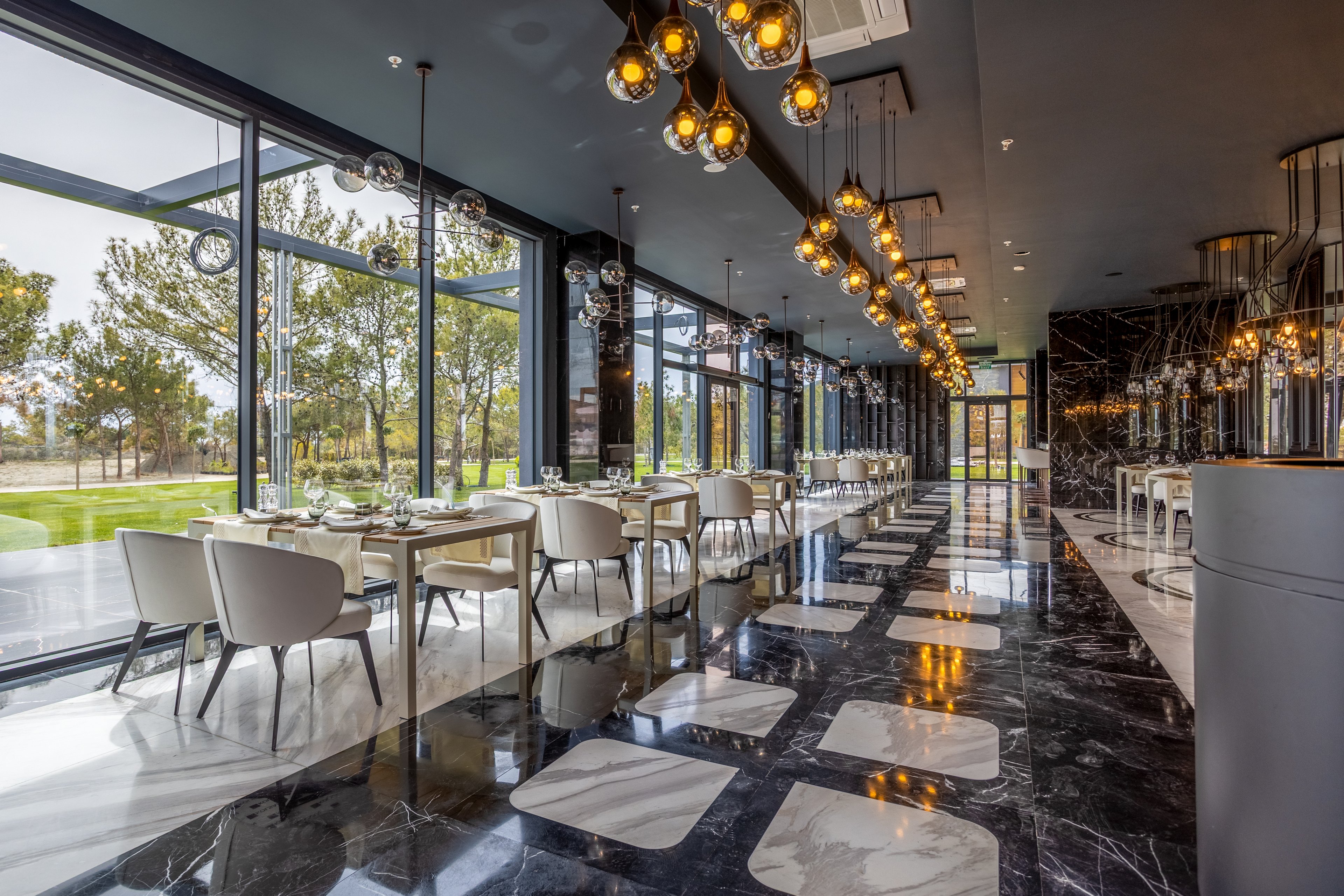 A contemporary restaurant interior features neatly arranged dining tables and white chairs along a glossy black marble floor. Large floor-to-ceiling windows reveal a green outdoor landscape with trees and lawn. Warm pendant lights hang from the ceiling, casting reflections on the polished surfaces and creating a sophisticated ambiance. The space appears ready for guests, with table settings prepared in a clean, minimalist style.