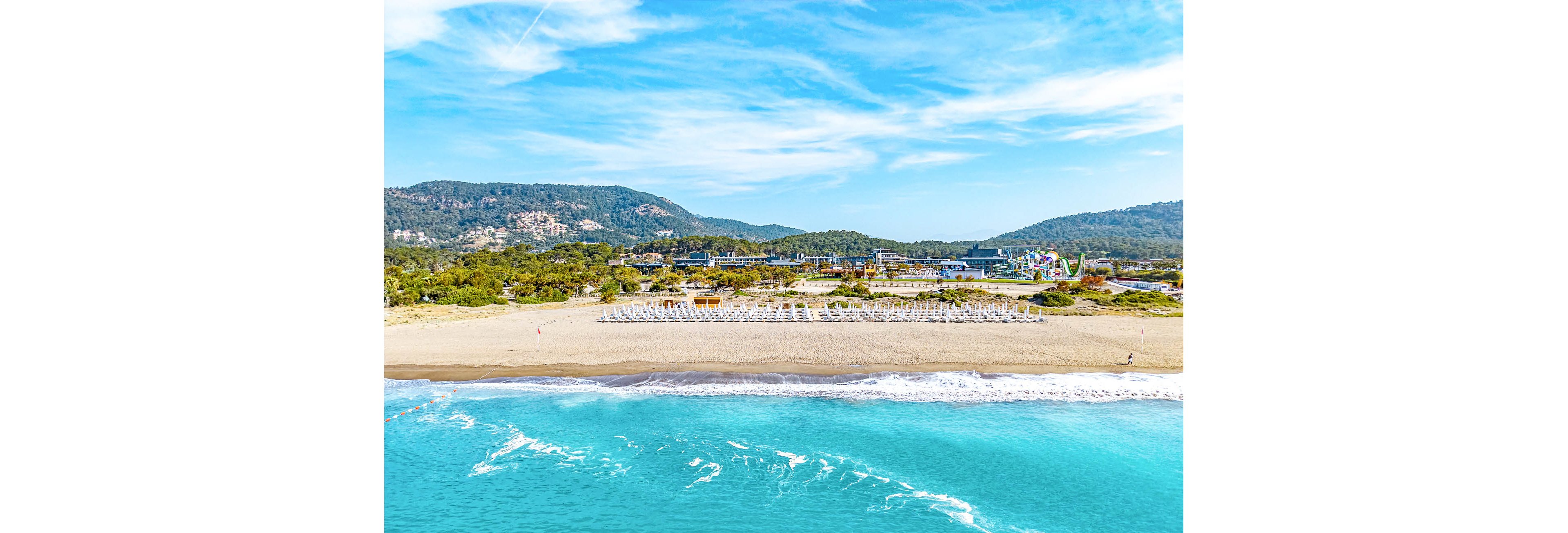 An aerial view captures a bright turquoise sea gently washing onto a wide sandy beach. Rows of neatly arranged sun loungers and umbrellas line the shore, indicating a well-organized resort area. Behind the beach, lush green vegetation and low hills stretch into the distance under a partly cloudy blue sky. The scene feels calm and inviting, highlighting a sunny coastal vacation destination.