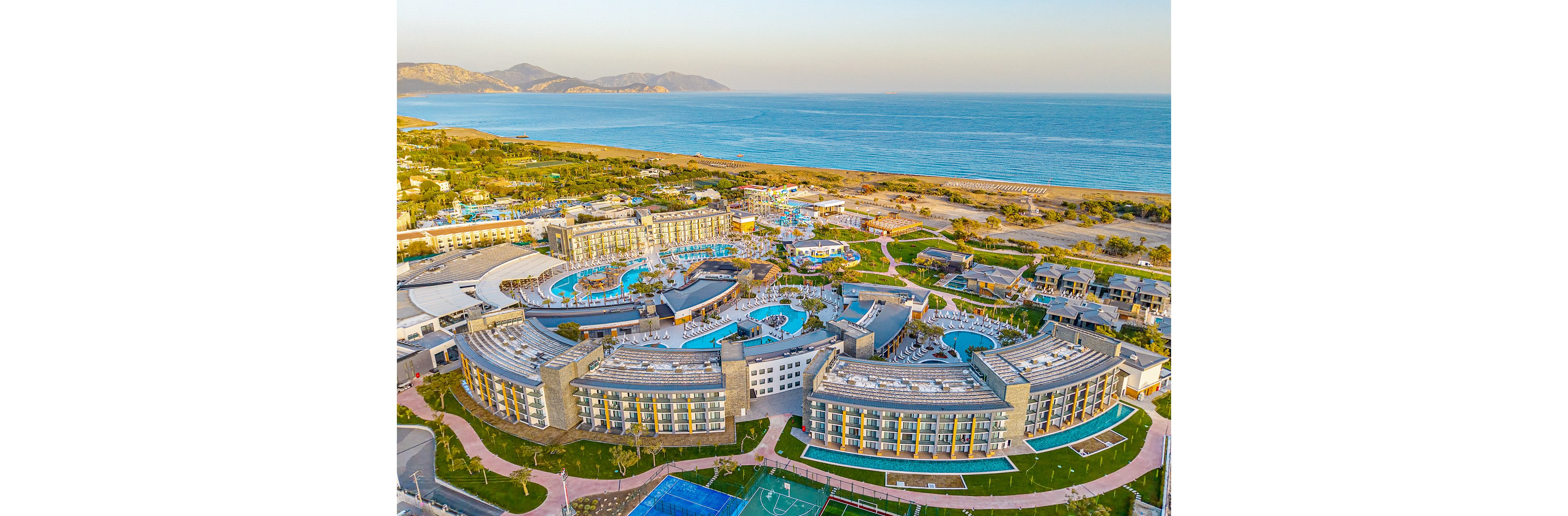 A large seaside resort complex is captured from an elevated aerial perspective. Curved multi-story hotel buildings surround several outdoor swimming pools and landscaped gardens. Sports courts and recreational areas are visible in the foreground, while the shoreline and calm sea stretch across the background. The scene is bathed in warm daylight, suggesting a relaxed vacation atmosphere.
