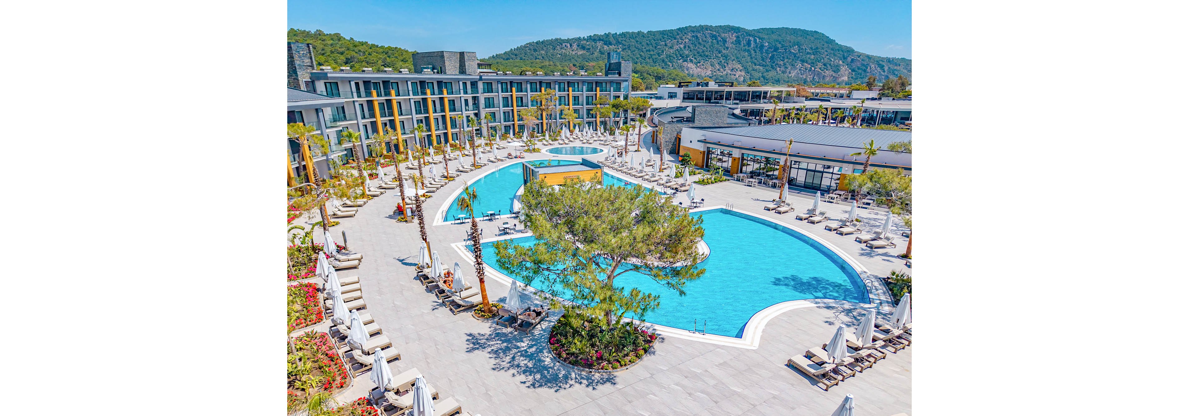 A large, curved swimming pool dominates the courtyard of a modern resort complex. Numerous white sun loungers and umbrellas surround the pool, creating a relaxed vacation atmosphere. Low-rise contemporary buildings frame the scene, with balconies overlooking the water. In the background, green hills and trees add a natural contrast to the bright blue pool and clear sky.