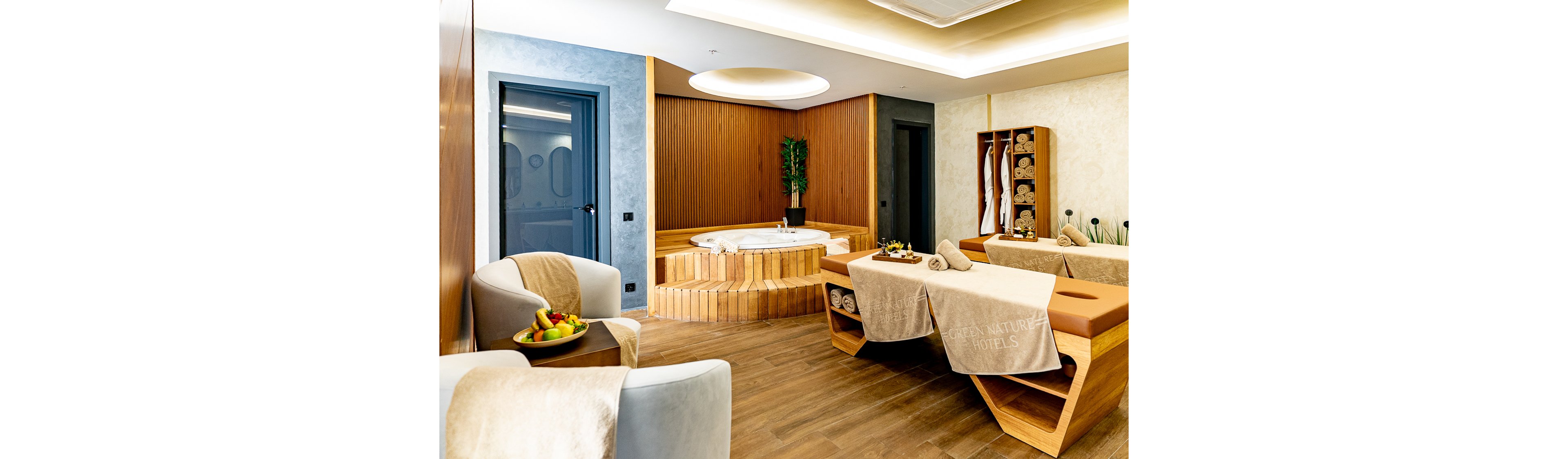 A contemporary spa treatment room features two massage tables covered with beige towels. In the background, a round jacuzzi tub is surrounded by warm wooden paneling and a tall green plant. Shelves hold neatly rolled towels and decor, while armchairs with small tables and fruit bowls create a welcoming seating area. Soft lighting and neutral tones contribute to a calm, luxurious atmosphere.
