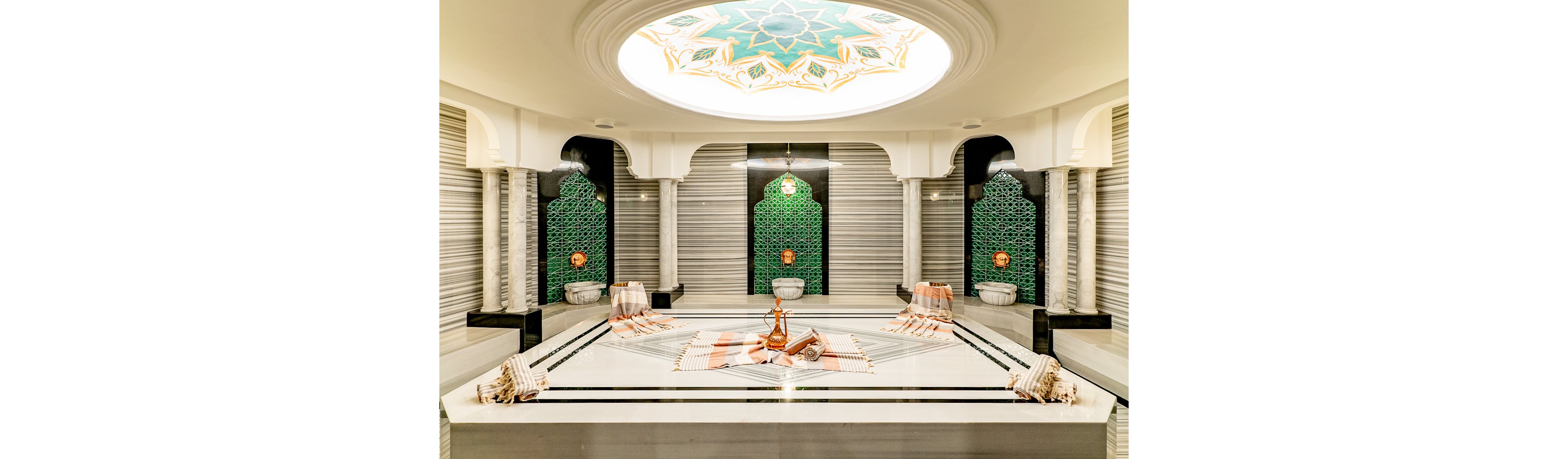 A luxurious Turkish hammam interior features a large central marble platform with copper vessels and neatly folded towels. The room is framed by ornate arches, green patterned wall niches, and decorative basins along the sides. Warm pendant lights and a detailed circular ceiling design create a bright yet soothing atmosphere. The overall scene emphasizes relaxation, cultural heritage, and spa-like comfort.