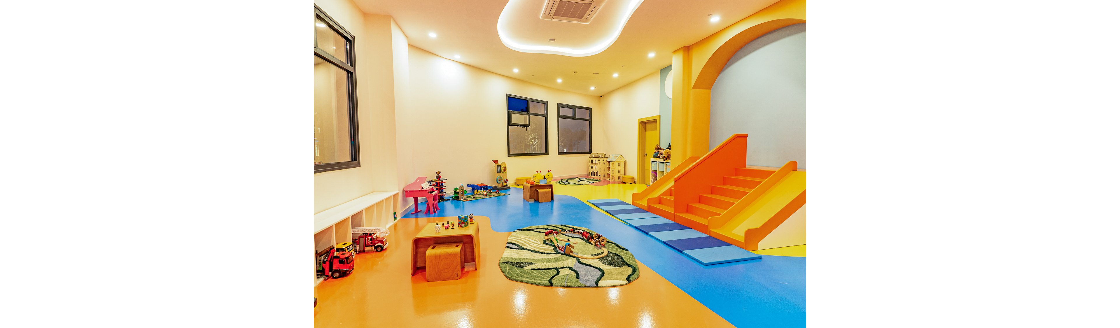 A spacious indoor playroom features a bright orange slide leading down to padded blue mats. The room is filled with colorful toys, low tables, and playful rugs arranged on a glossy blue and orange floor. Soft, warm lighting and curved ceiling details create a welcoming atmosphere for children. Large windows and neatly organized storage cubbies enhance the clean, modern design of the space.