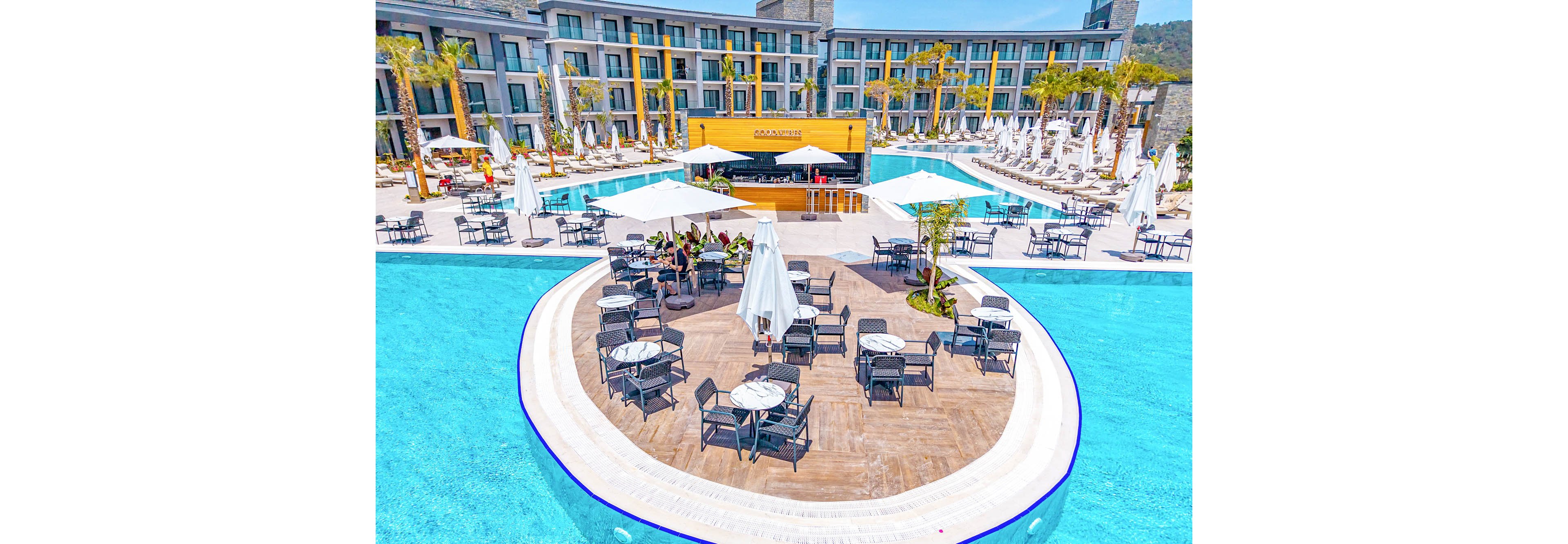 A large resort swimming pool features a circular island deck with tables, chairs, and umbrellas arranged for guests. The pool surrounds the island, creating a striking centerpiece in front of a modern multi-story hotel building. A bright yellow pool bar stands at the far side, with sun loungers and white umbrellas lining the poolside terrace. Palm trees and distant hills complete the sunny vacation atmosphere.