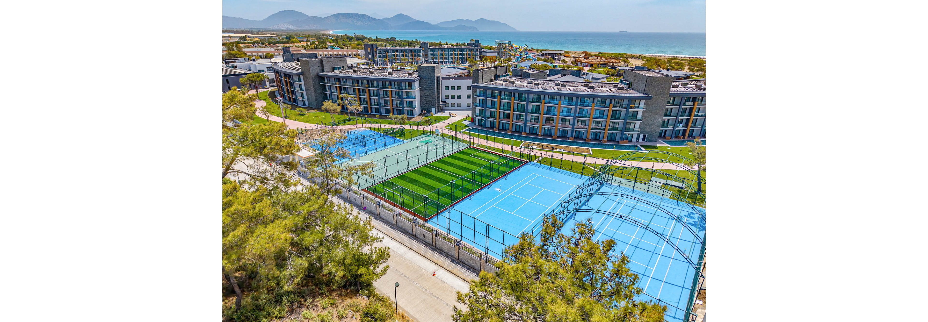 A modern coastal resort complex features multiple outdoor sports courts, including bright blue tennis courts and a green artificial turf field. The courts are surrounded by trees and walking paths, with contemporary multi-story buildings in the background. Beyond the resort, the sea and distant mountains create a scenic backdrop under a clear sky. The image highlights recreational facilities in a vacation setting.