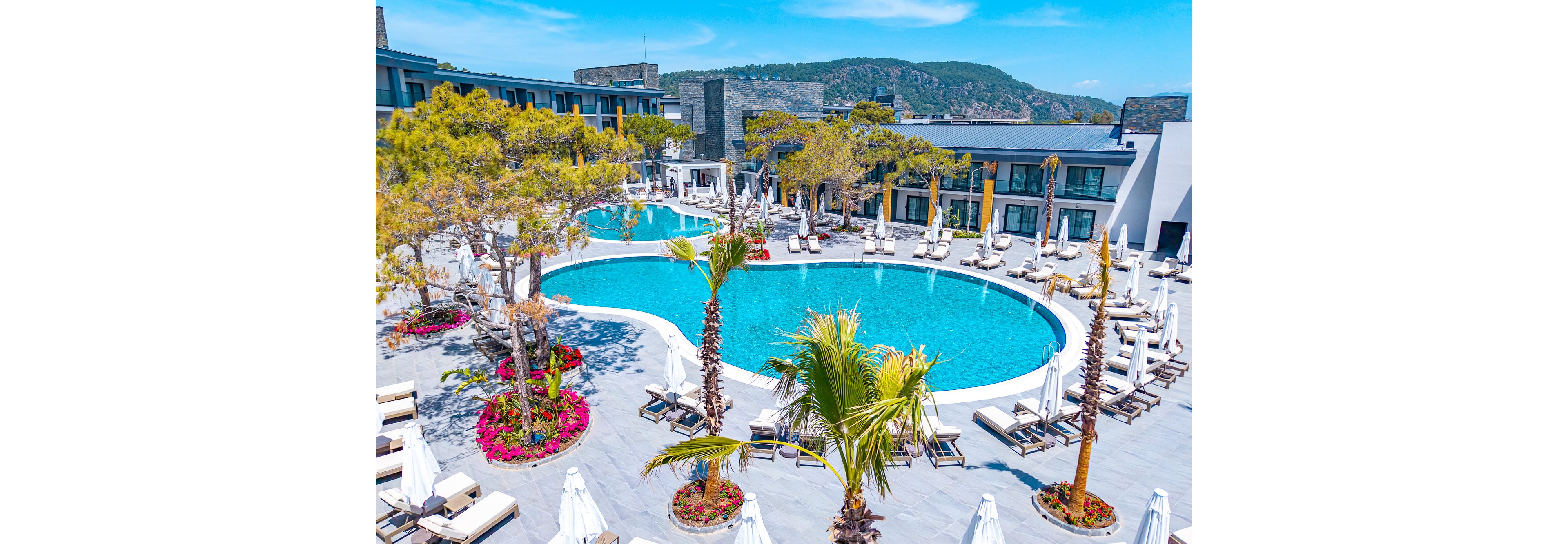 A large resort swimming pool curves through a modern hotel courtyard under a bright blue sky. Numerous white sun loungers and umbrellas surround the pool, creating a relaxing vacation atmosphere. Palm trees and colorful flower beds add tropical greenery and vibrant accents to the scene. Low-rise contemporary buildings and distant hills frame the tranquil outdoor setting.