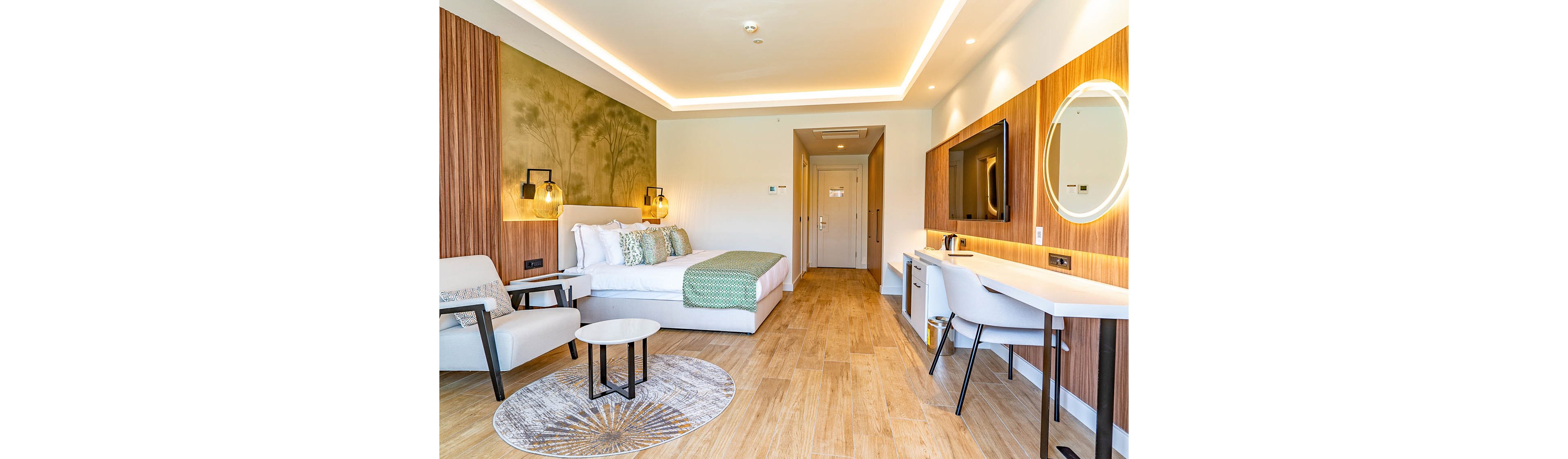 A contemporary hotel room features a neatly made double bed with green and white textiles. The space includes a comfortable armchair and side table near a round rug, creating a small seating area. Warm wooden wall panels and flooring complement recessed ceiling lighting and hanging bedside lamps. A long white desk with chair, wall-mounted television, and illuminated round mirror complete the stylish interior.
