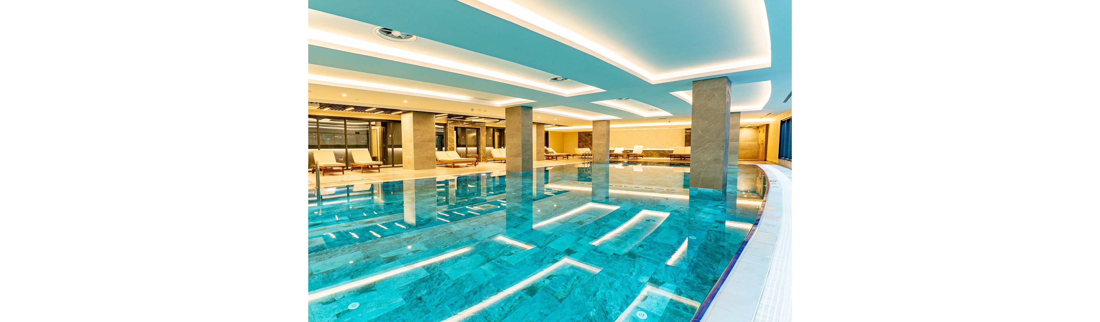 A spacious indoor swimming pool dominates a contemporary spa or hotel setting. The turquoise water reflects the bright recessed ceiling lights and clean architectural lines. Lounge chairs and seating areas are arranged along the far side, suggesting a relaxing wellness environment. Large windows and glass doors add openness, while neutral tones contrast with the vivid blue pool.